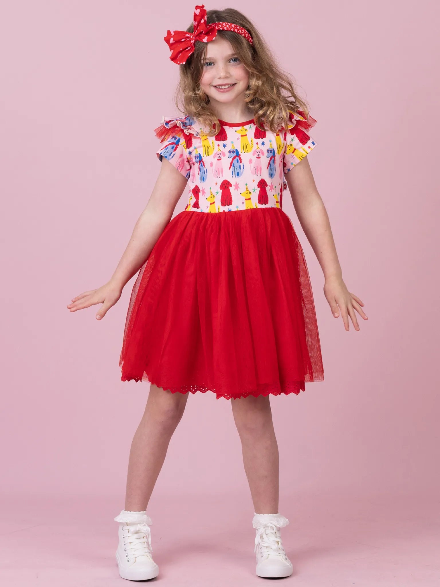 A young girl modeling a red and white dress with a colorful patterned bodice, white tulle skirt, and a red bow headband, standing against a solid pink background.