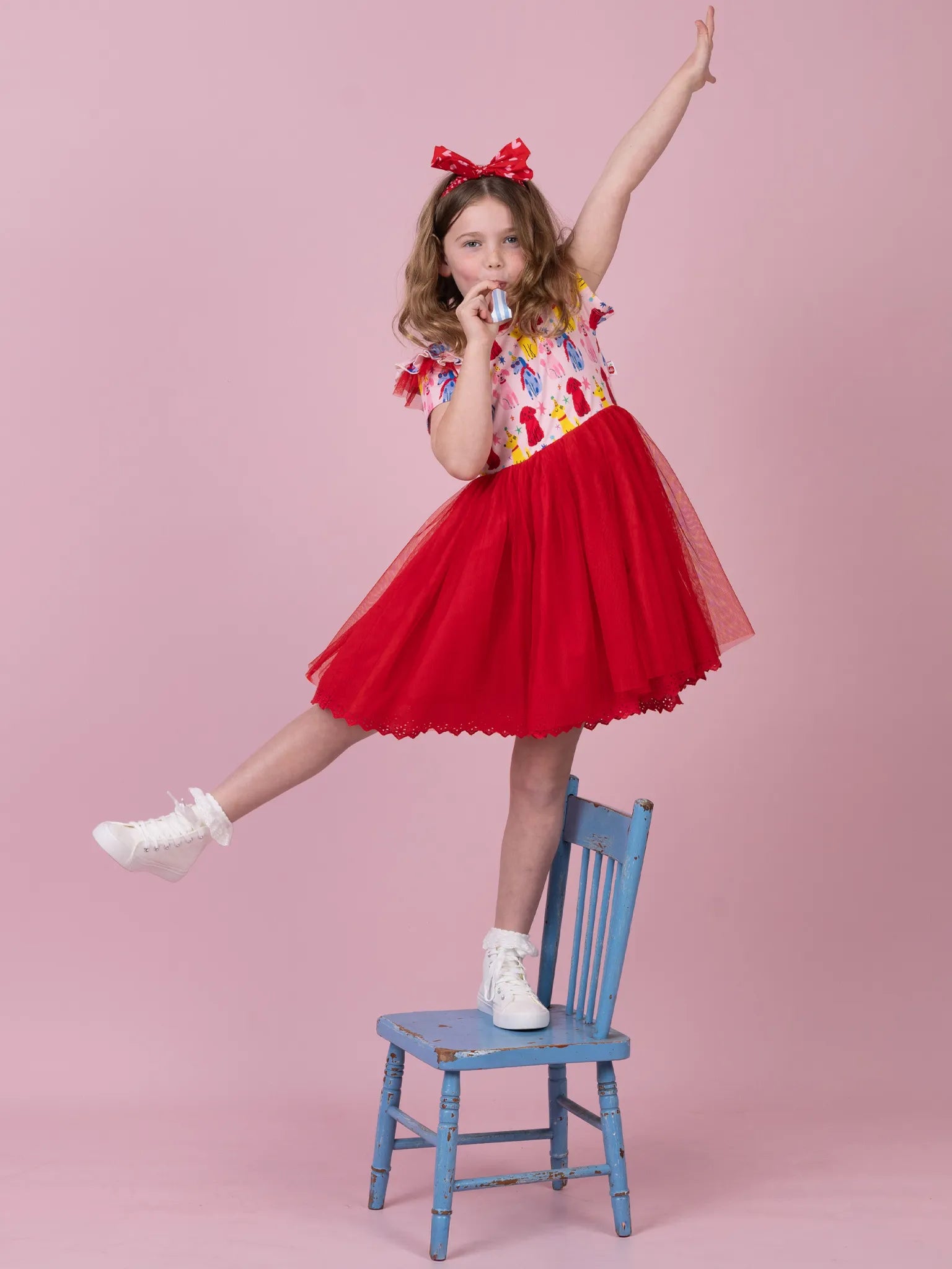 A young girl wearing a red tutu dress with a colorful patterned bodice and a red bow in her hair, posing on a small blue chair against a solid pink background.