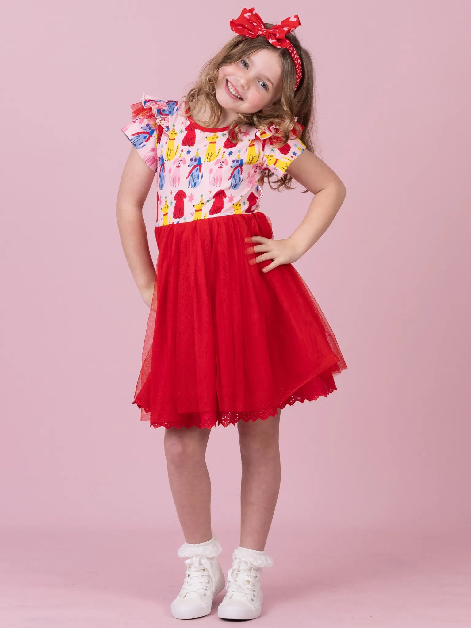 A young girl modeling a short-sleeve dress with a colorful ice cream cone print bodice and a red tulle skirt, paired with a red bow headband and white sneakers, against a solid pink background.