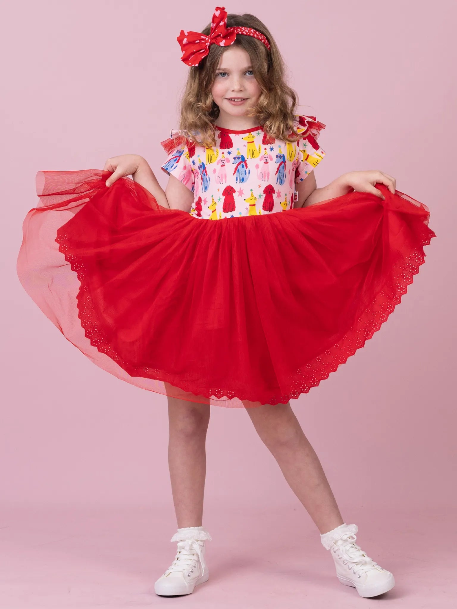 A young girl modeling a red tulle tutu skirt dress with a colorful ice cream cone print bodice, holding the skirt outwards against a solid pink studio background.
