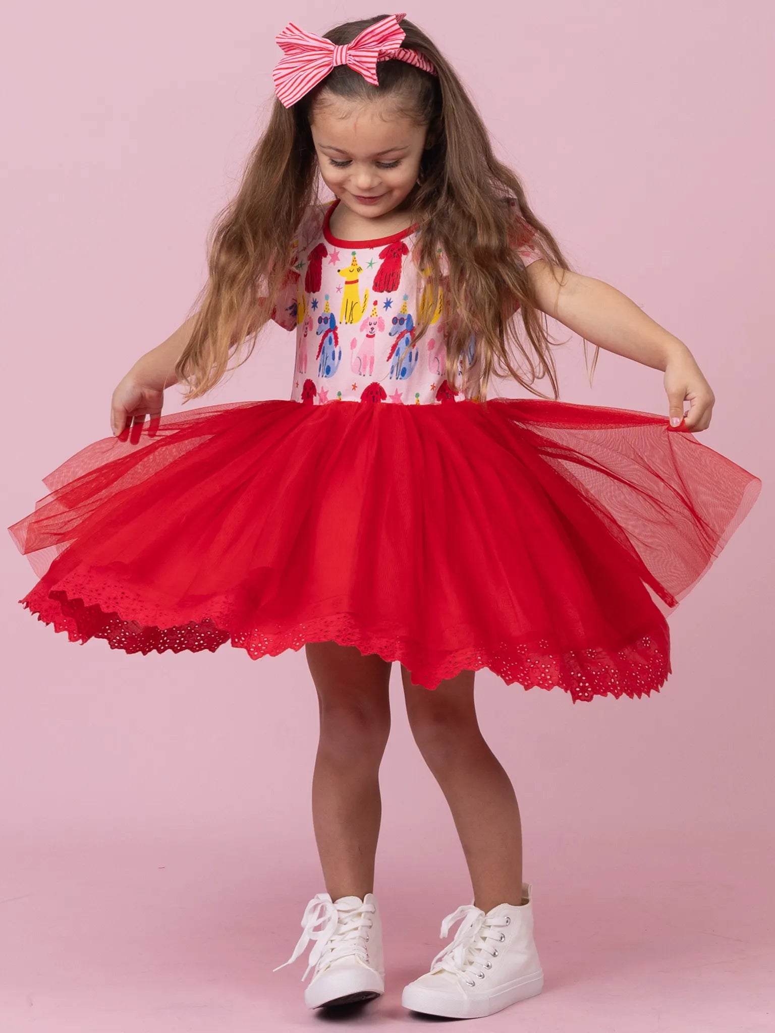 A young girl with long brown hair wearing a red tutu skirt, a colorful patterned top, and a pink and white striped bow headband, posing against a solid pink background.