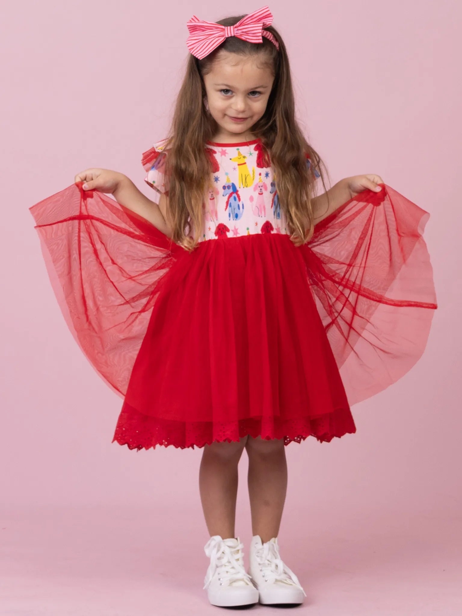 A young girl wearing a red and white dress with a colorful bodice, a red tulle skirt, and holding out her sheer red sleeves against a solid pink studio background. She is also wearing a pink and white bow headband and white sneakers.