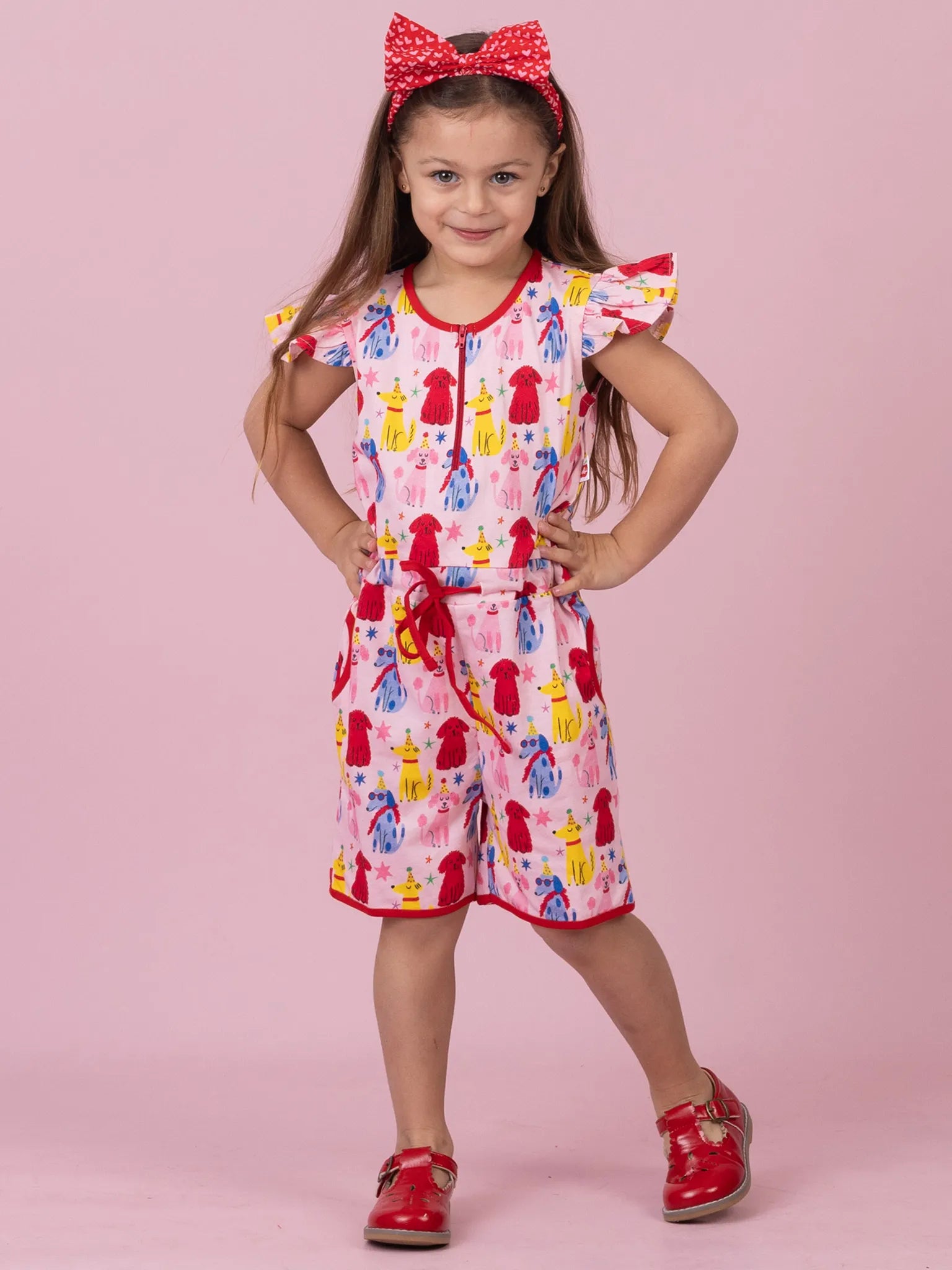 A young girl modeling a pink short-sleeve romper with a colorful red, yellow, and blue pattern, styled with a red polka-dot bow headband and red shoes against a solid pink background.
