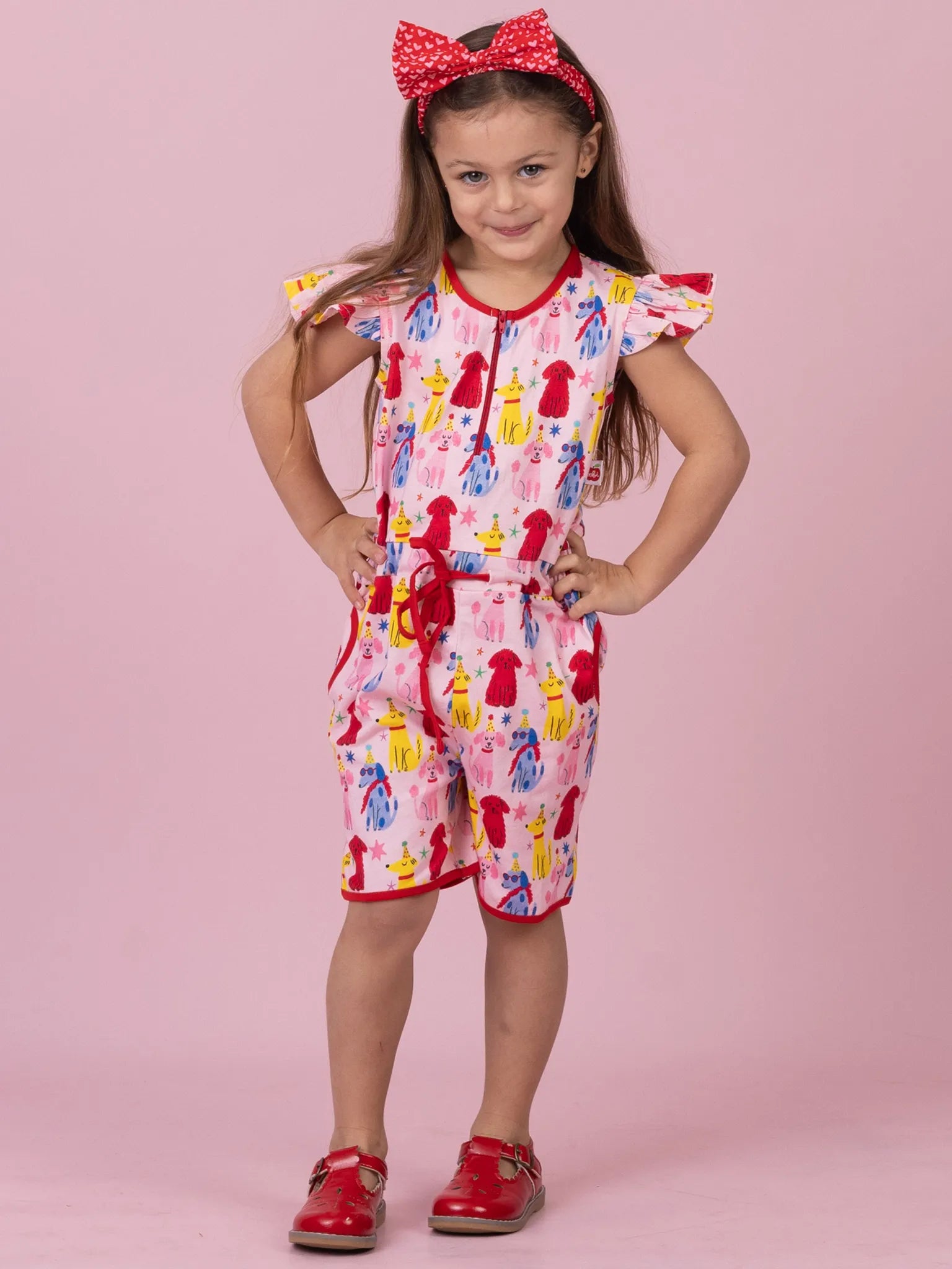 A young girl modeling a pink and red short-sleeve romper with a colorful fashion-dress pattern, a red bow headband, and red shoes, standing against a solid pink background.