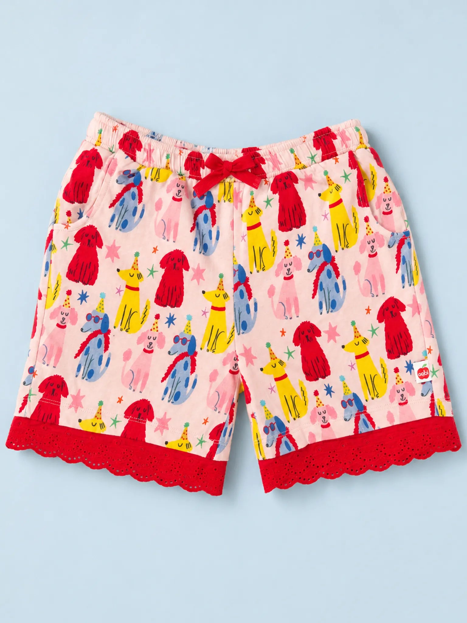 A pair of children's pajama shorts featuring a colorful cartoon dog print on a light pink background, with a red drawstring and red scalloped lace trim at the hem, shown flat on a light blue background.