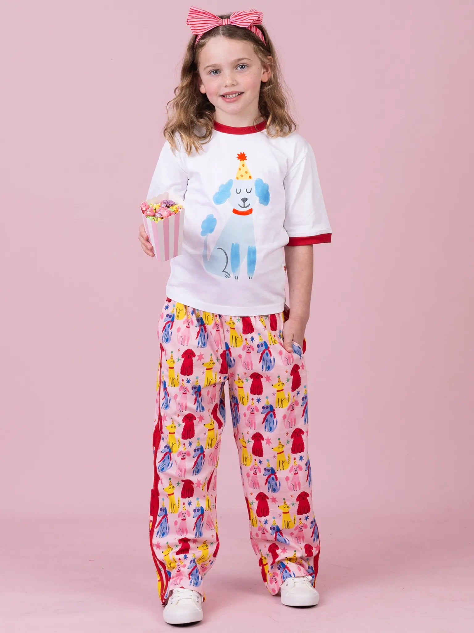 A young girl stands against a solid pink studio background wearing a white short-sleeve t-shirt with a light blue dog graphic and red trim, paired with pink patterned pants featuring a colorful ice cream cone print. She is holding a small pink cup and wearing a pink bow headband.