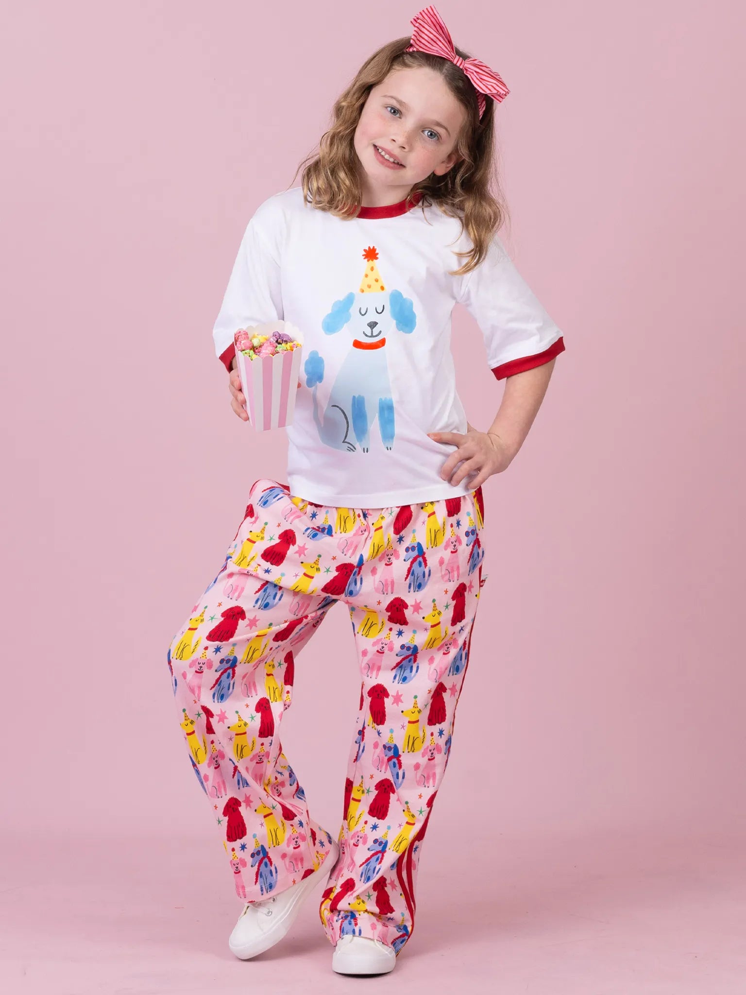 A young girl modeling a matching pajama set against a solid pink studio background. The set includes a white short-sleeve top with red trim and a blue dog graphic, paired with matching pink pants covered in a colorful bird print.