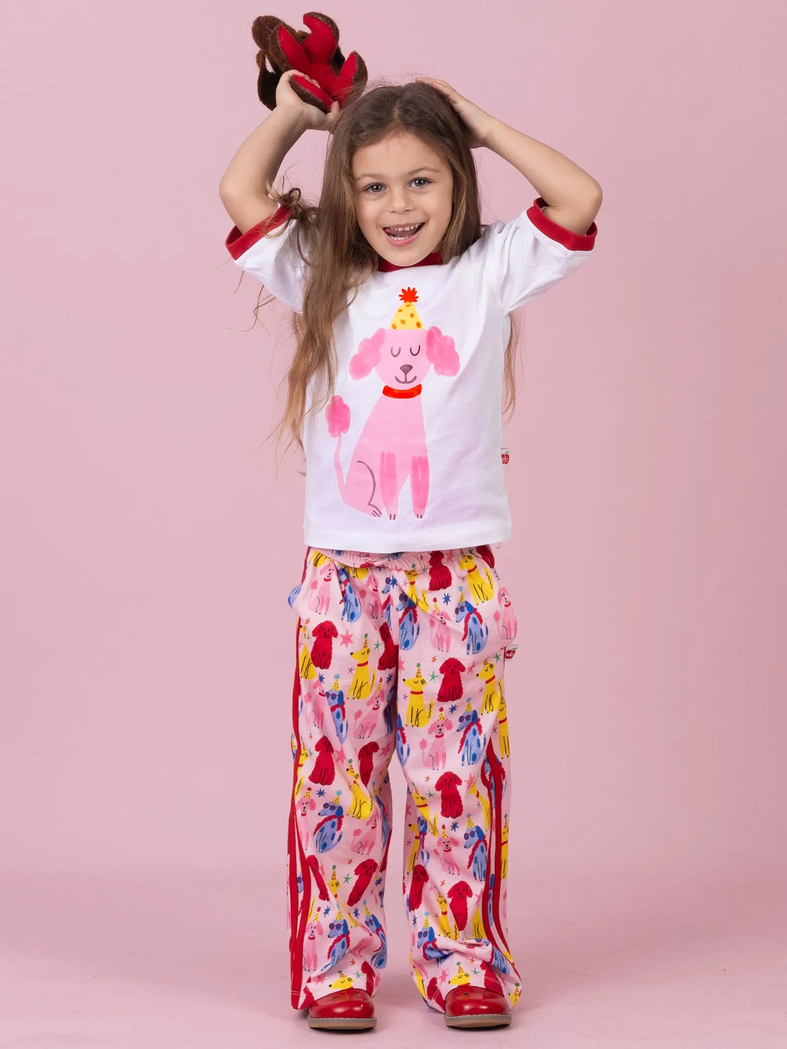 A young girl modeling a two-piece pajama set consisting of a white short-sleeve t-shirt with a pink poodle graphic and red cuffs, paired with matching pink patterned pants. She is posing against a solid pink studio background.