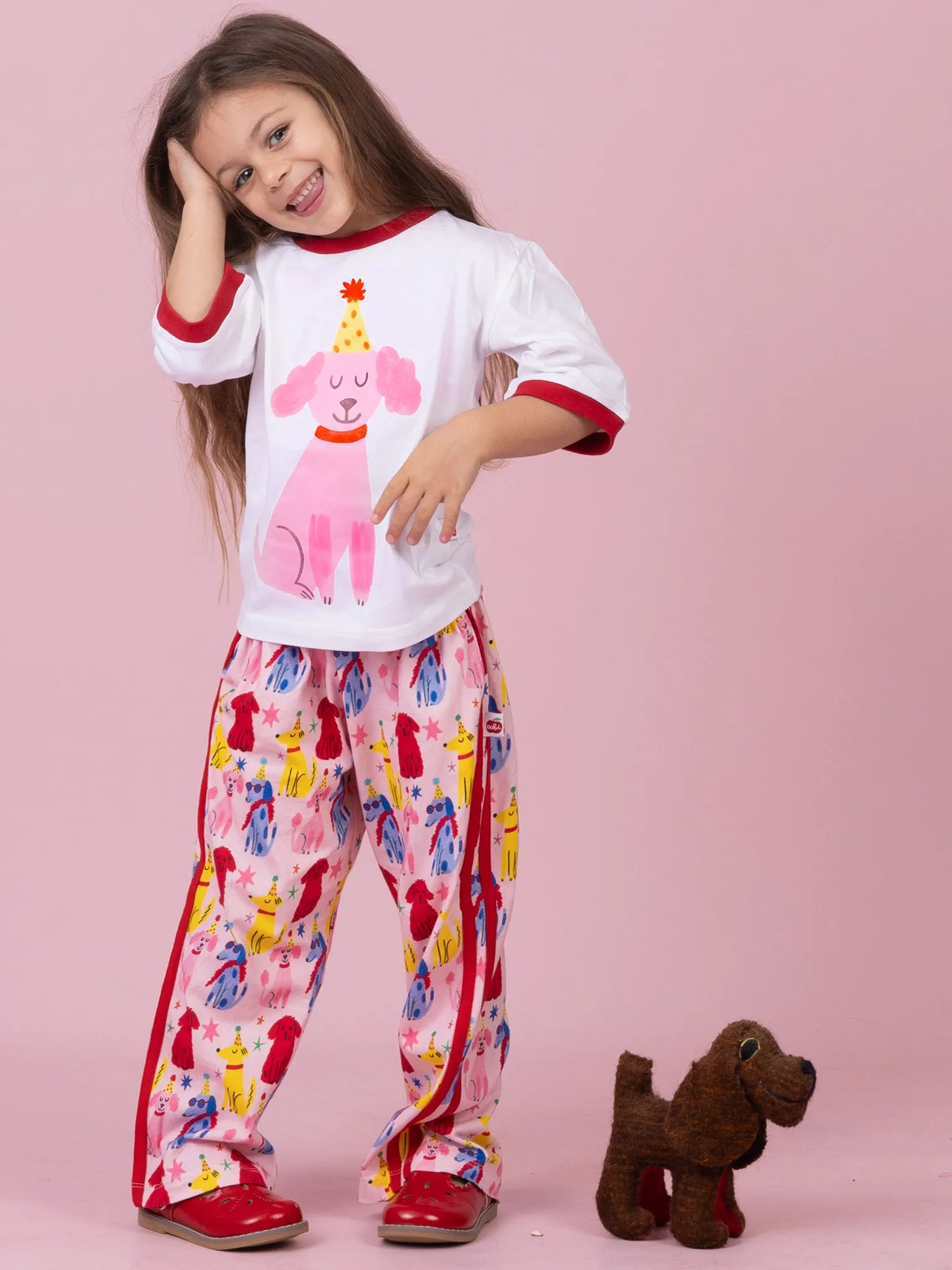 A young girl modeling a two-piece pajama set consisting of a white short-sleeve top with red trim and a pink dog graphic, paired with matching pink pants featuring a colorful dog print and red side stripes. She is standing against a solid pink background next to a small brown plush dog toy.