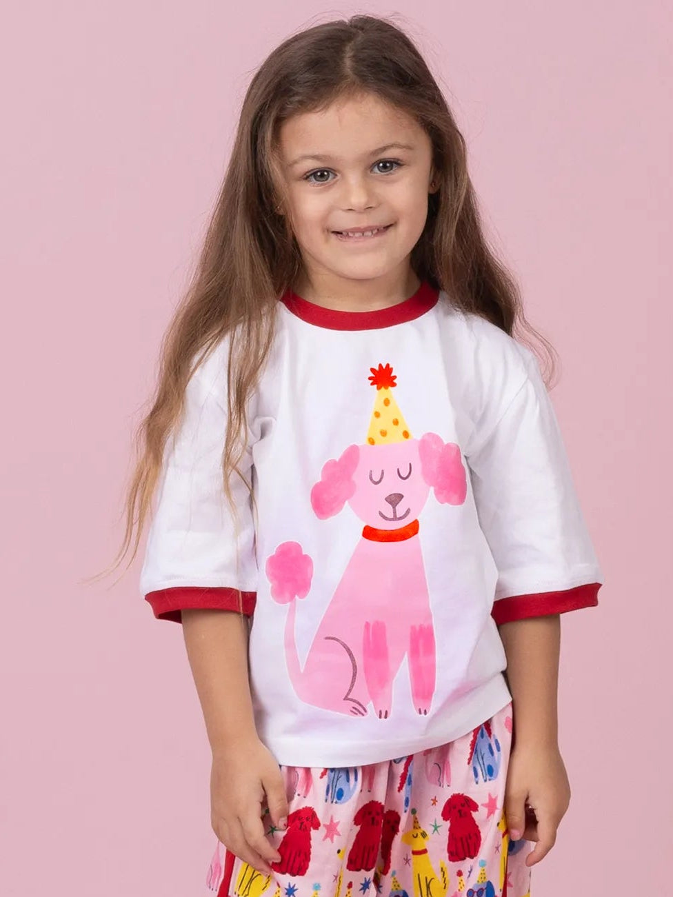 A young girl stands against a solid pink studio background wearing a white short-sleeve pajama top with red trim and a pink poodle graphic, paired with matching pink patterned pajama pants. A small brown plush dog toy sits on the floor beside her.