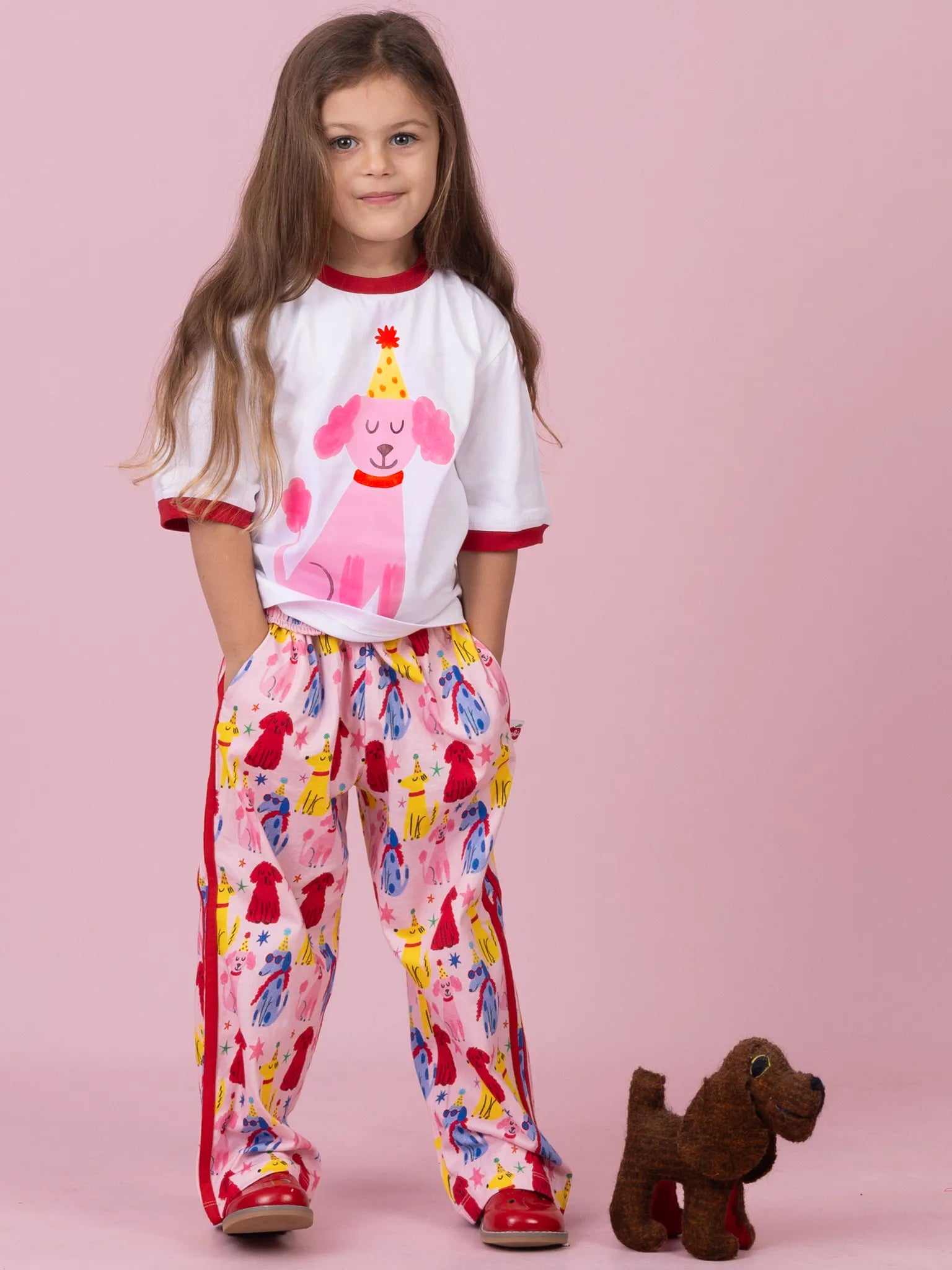 A young girl stands against a solid pink background wearing a matching pajama set consisting of a white short-sleeve top with red trim and a pink dog graphic, paired with pink pants featuring a colorful dog print. A small brown plush dog toy sits next to her.