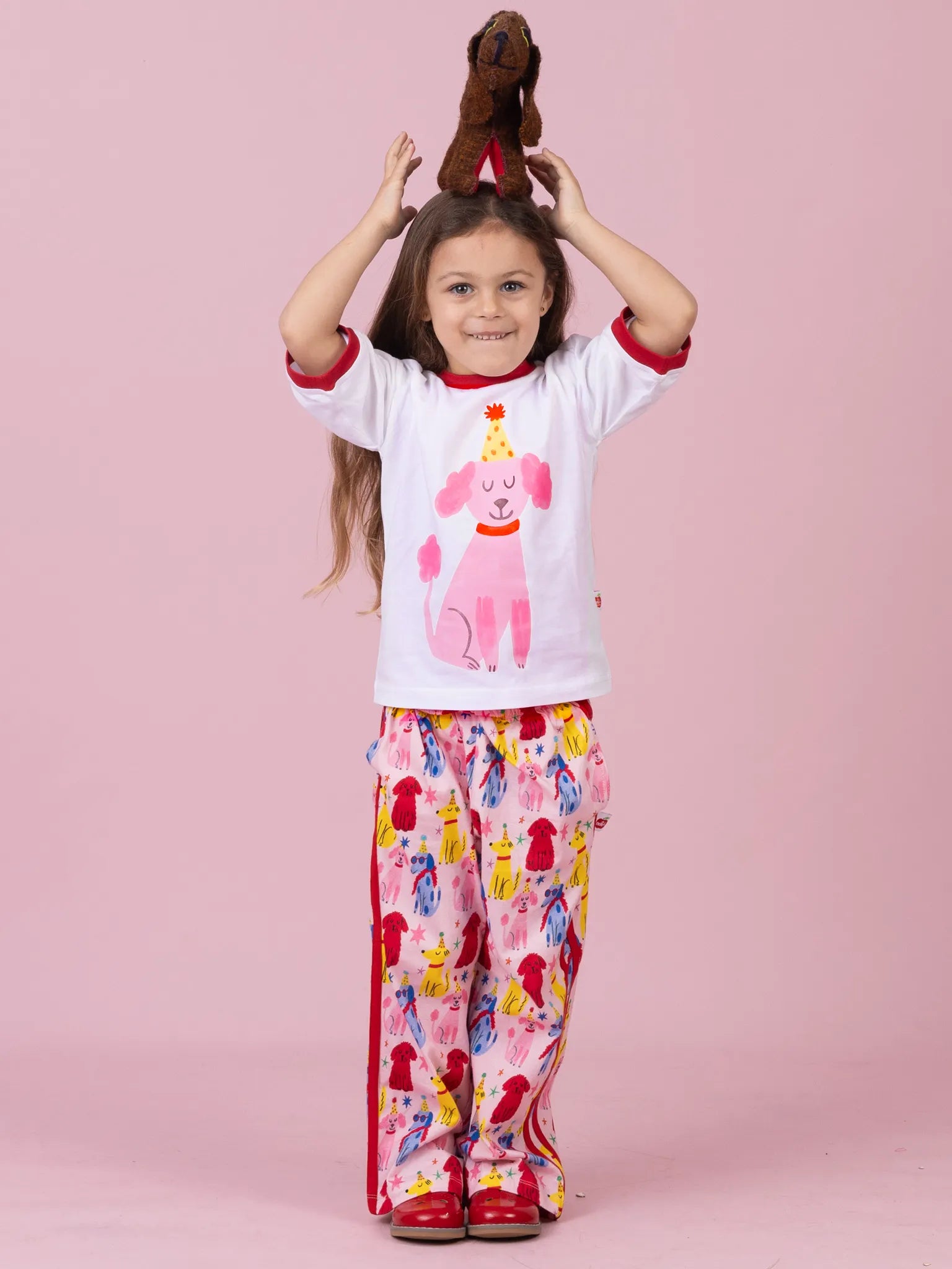 A young girl modeling a white short-sleeve pajama top with a pink dog graphic and red trim, paired with pink patterned pajama pants, standing against a solid pink background.