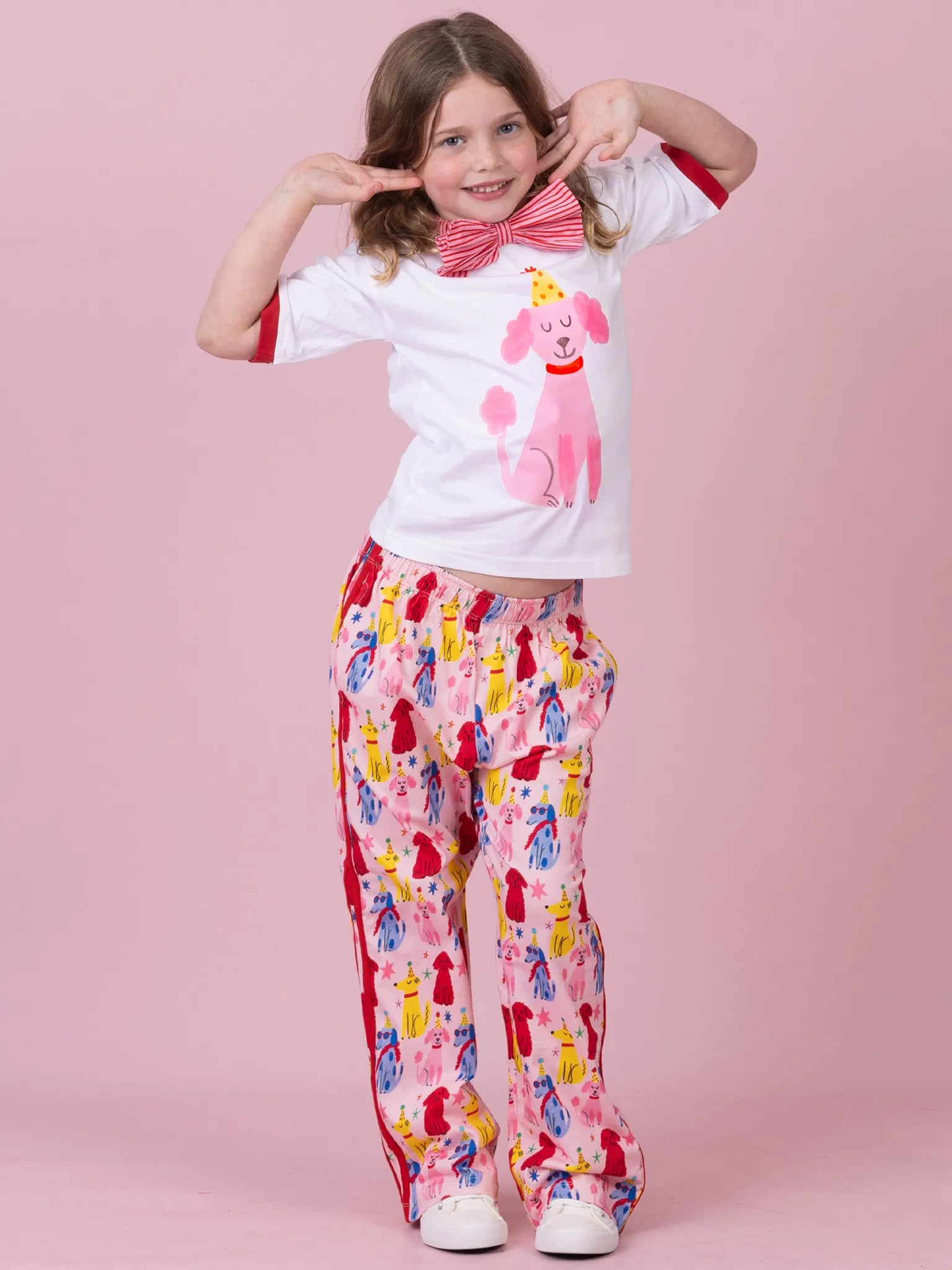 A young girl modeling a two-piece pajama set consisting of a white short-sleeve t-shirt with a pink poodle and bow tie graphic, and matching pink pants with a colorful poodle print, set against a solid pink background.