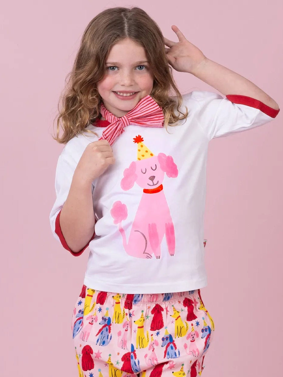 A young girl modeling a two-piece pajama set consisting of a white short-sleeve top with red trim and a pink poodle graphic, paired with matching pink patterned pants featuring red side stripes, set against a solid pink background.