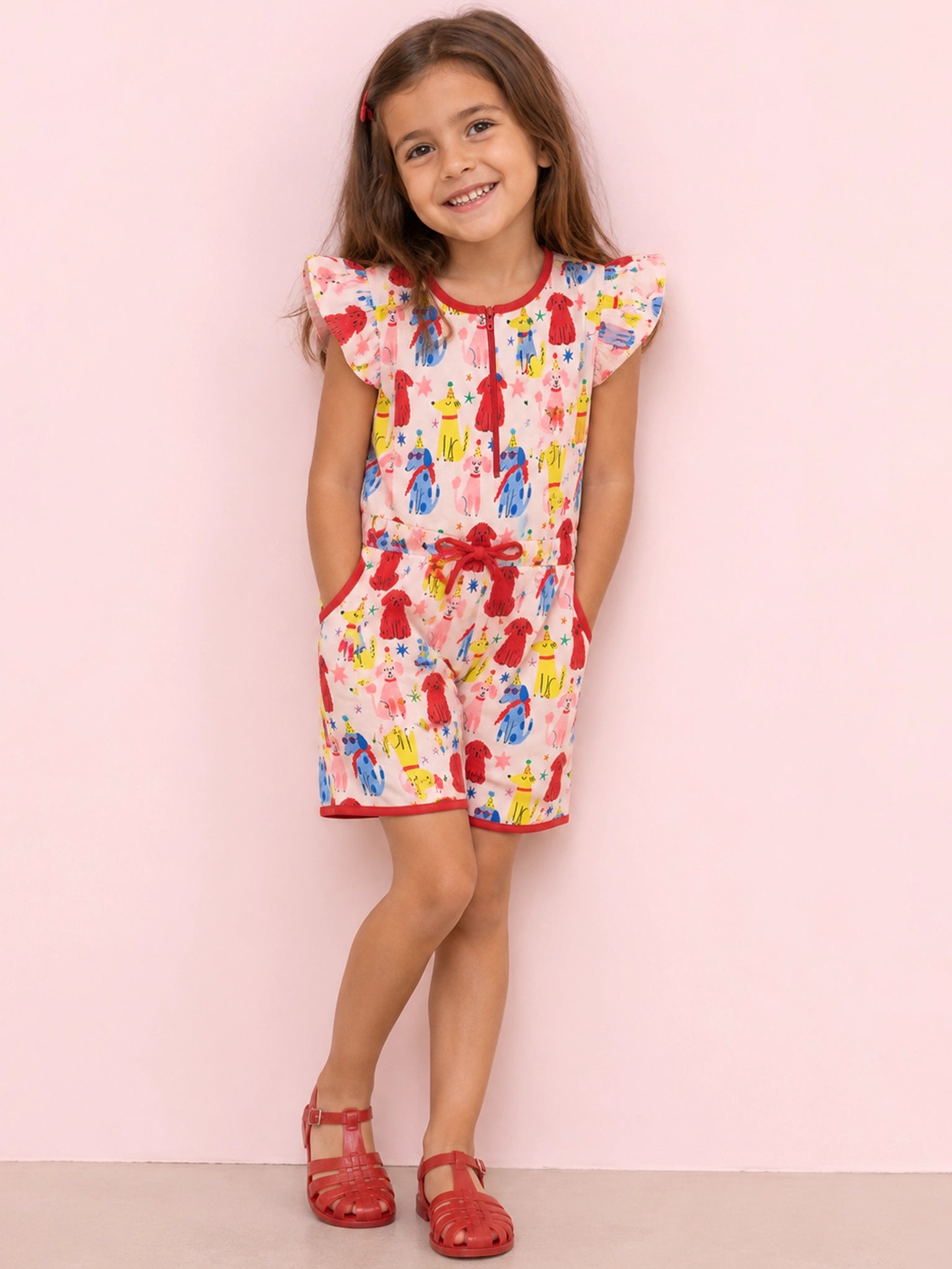 A young girl with brown hair stands against a light pink wall wearing a short white romper with a colorful painterly print, red trim, and red sandals.