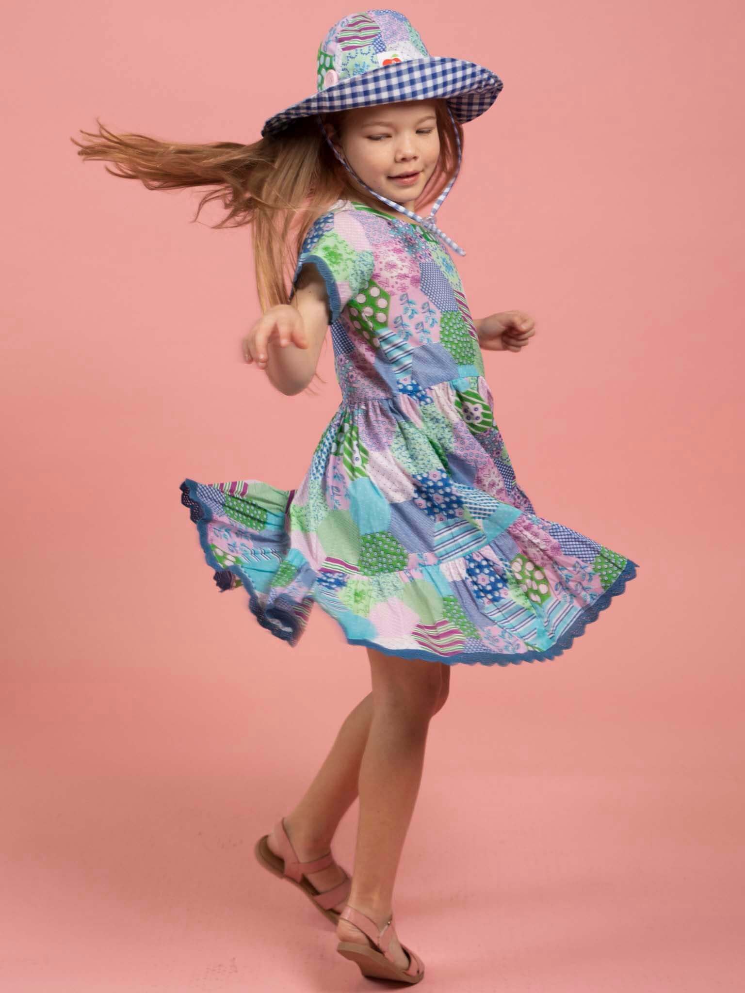 Child wearing a colorful patchwork dress and checkered hat on a pink background