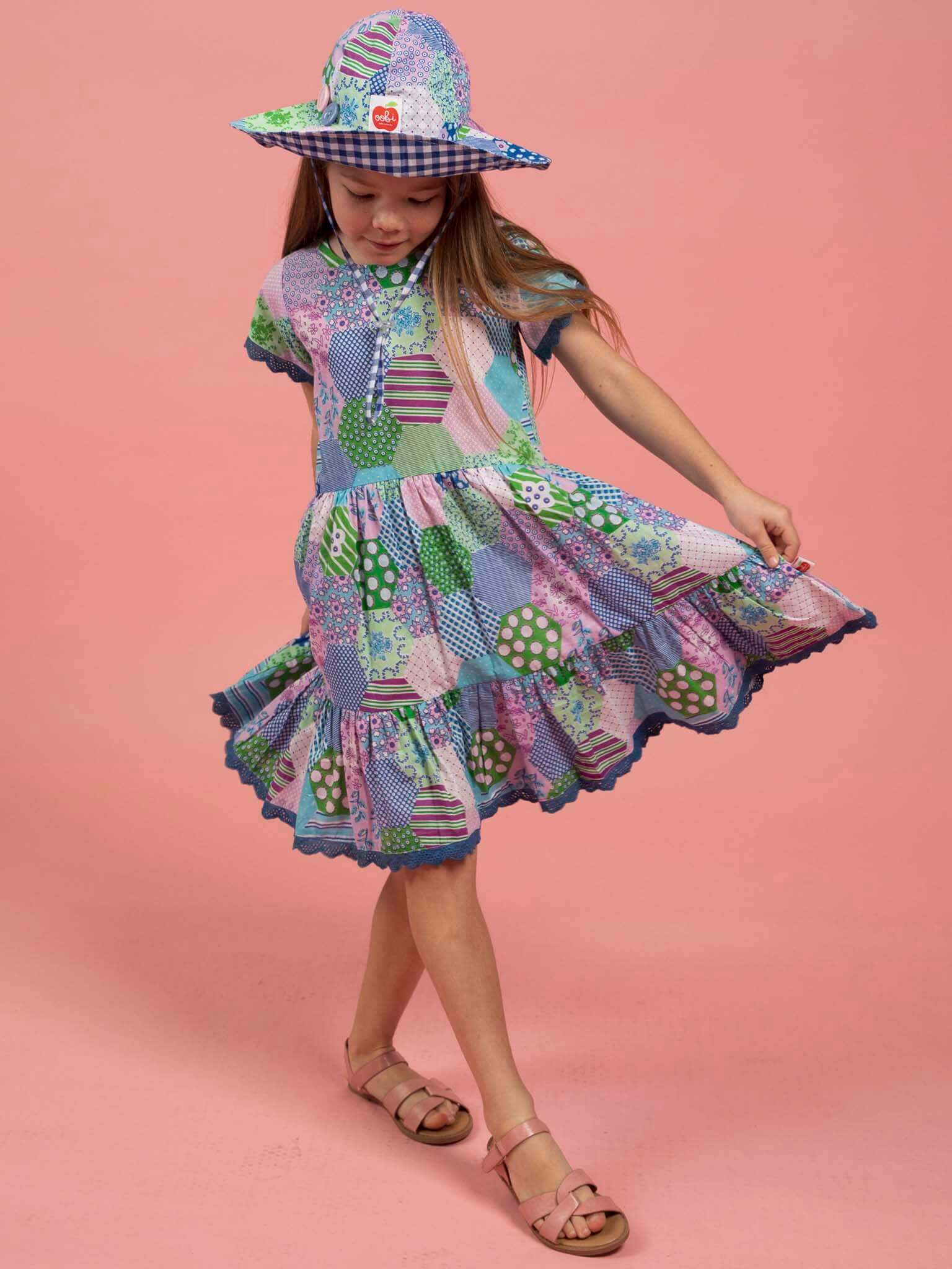 Young girl wearing a colorful patchwork dress and hat on a pink background