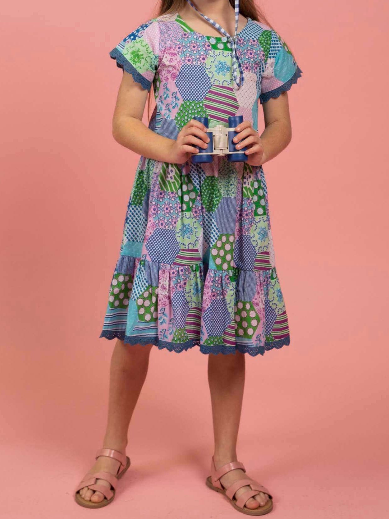 Young girl wearing a colorful patchwork dress and checkered hat on a pink background