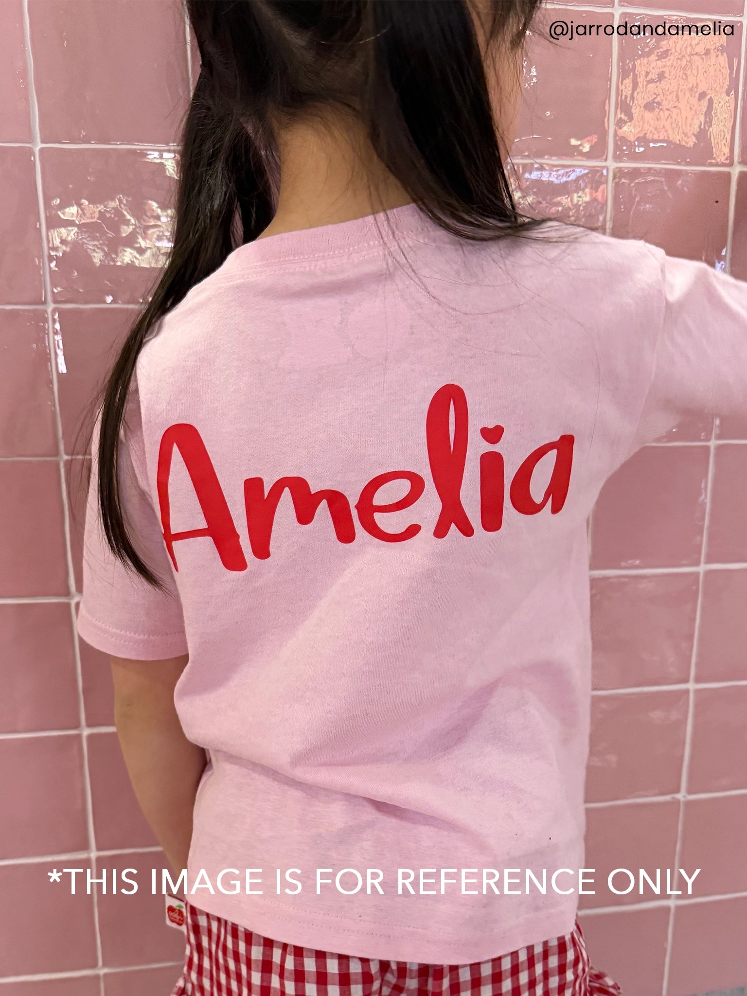 A child shown from the back wearing a light pink T-shirt with the name 'Amelia' printed in red script across the back, standing against a pink tiled wall. A watermark and reference text are visible on the image.