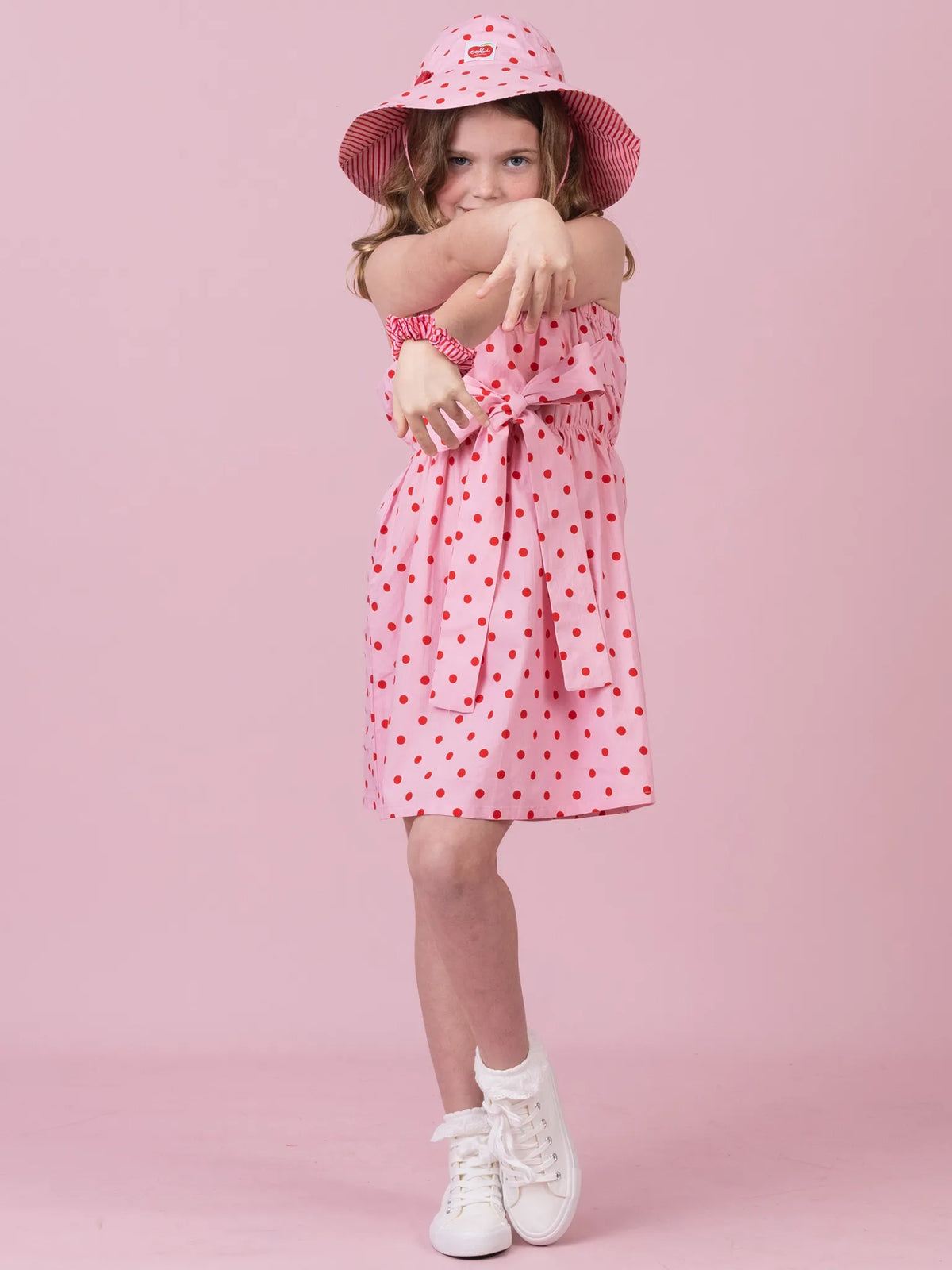 A young girl wearing a pink and red polka dot dress with a matching hat and white sneakers, posing against a solid pink studio background.