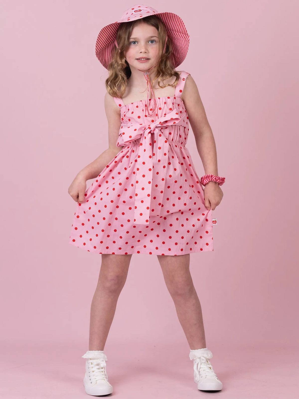 A young girl modeling a pink sleeveless dress with red polka dots, a matching pink hat, and white sneakers against a solid pink background.