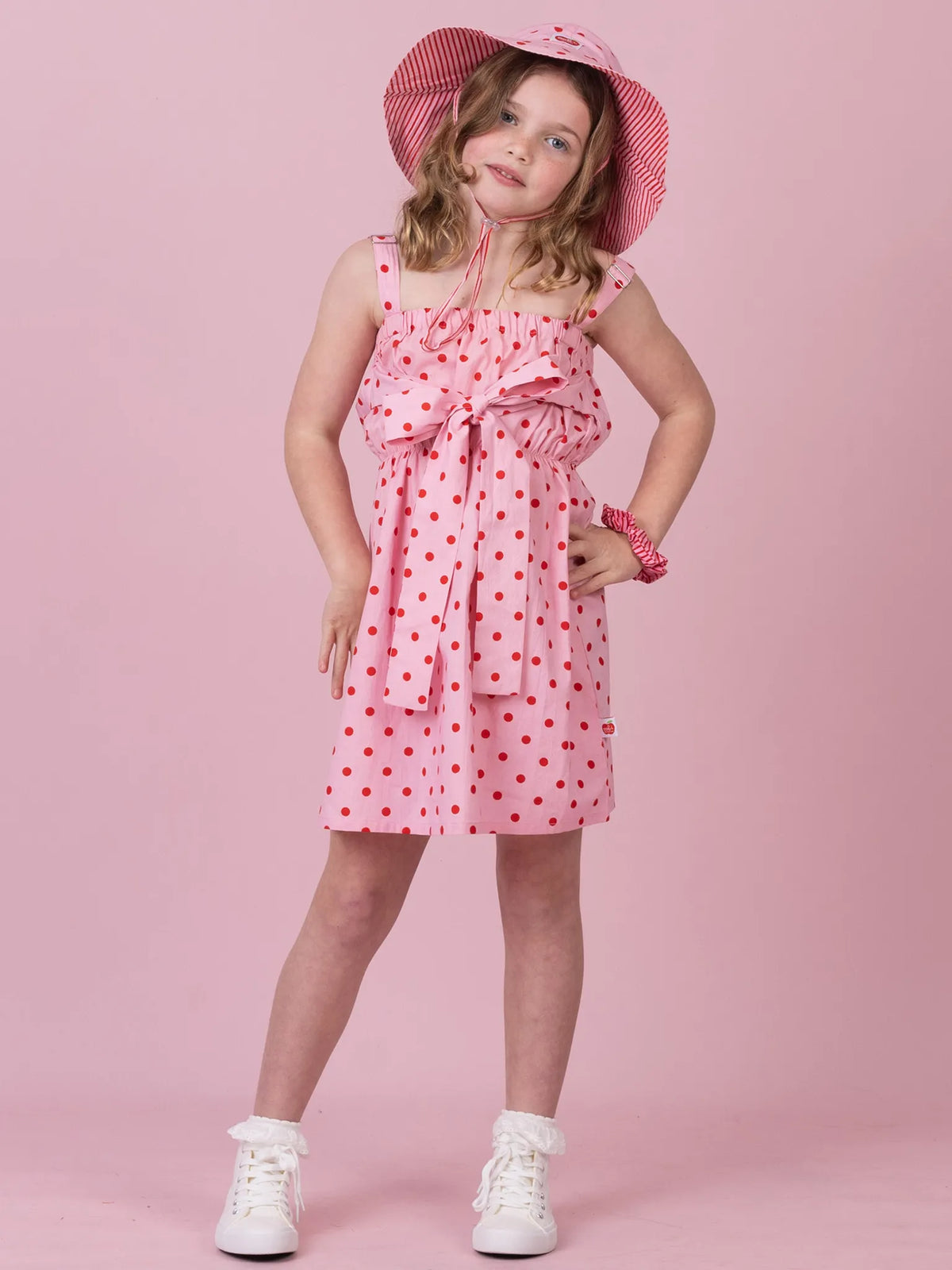 A young girl modeling a pink sleeveless dress with red polka dots, a matching pink hat, and white sneakers against a solid pink background.