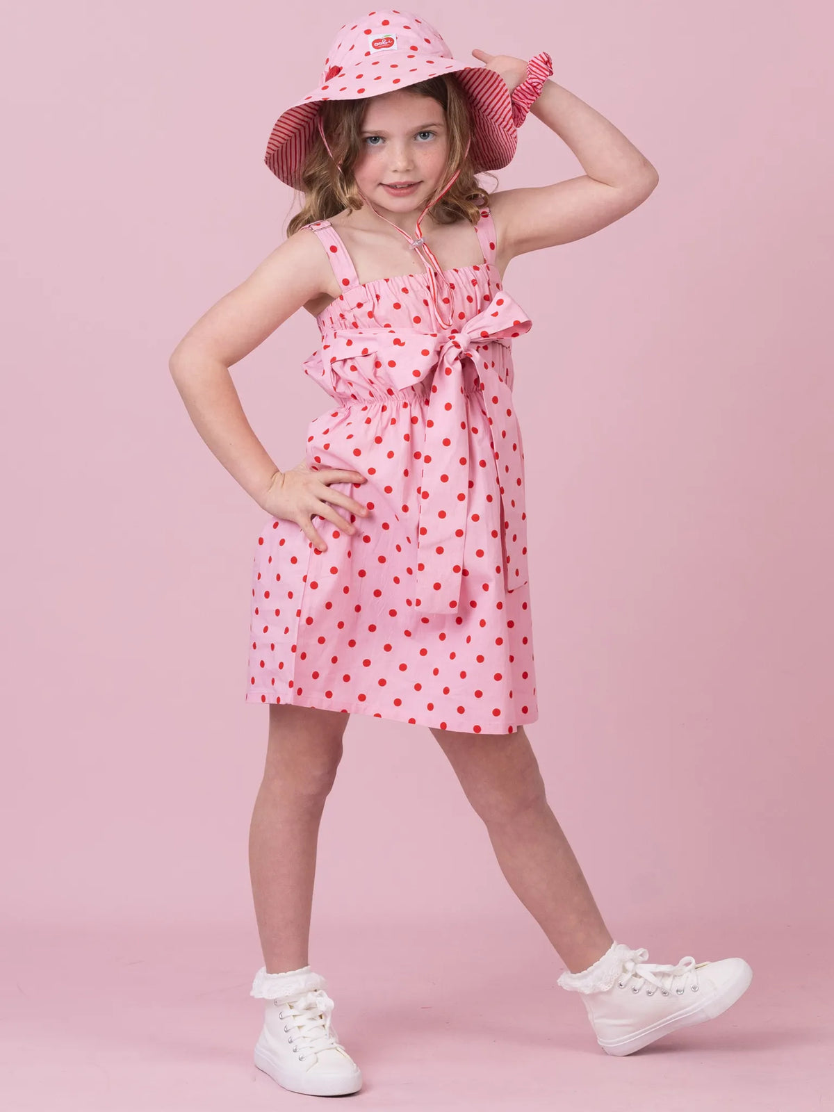 A young girl modeling a pink sleeveless dress with red polka dots, a matching pink sun hat, and white sneakers against a solid pink studio background.