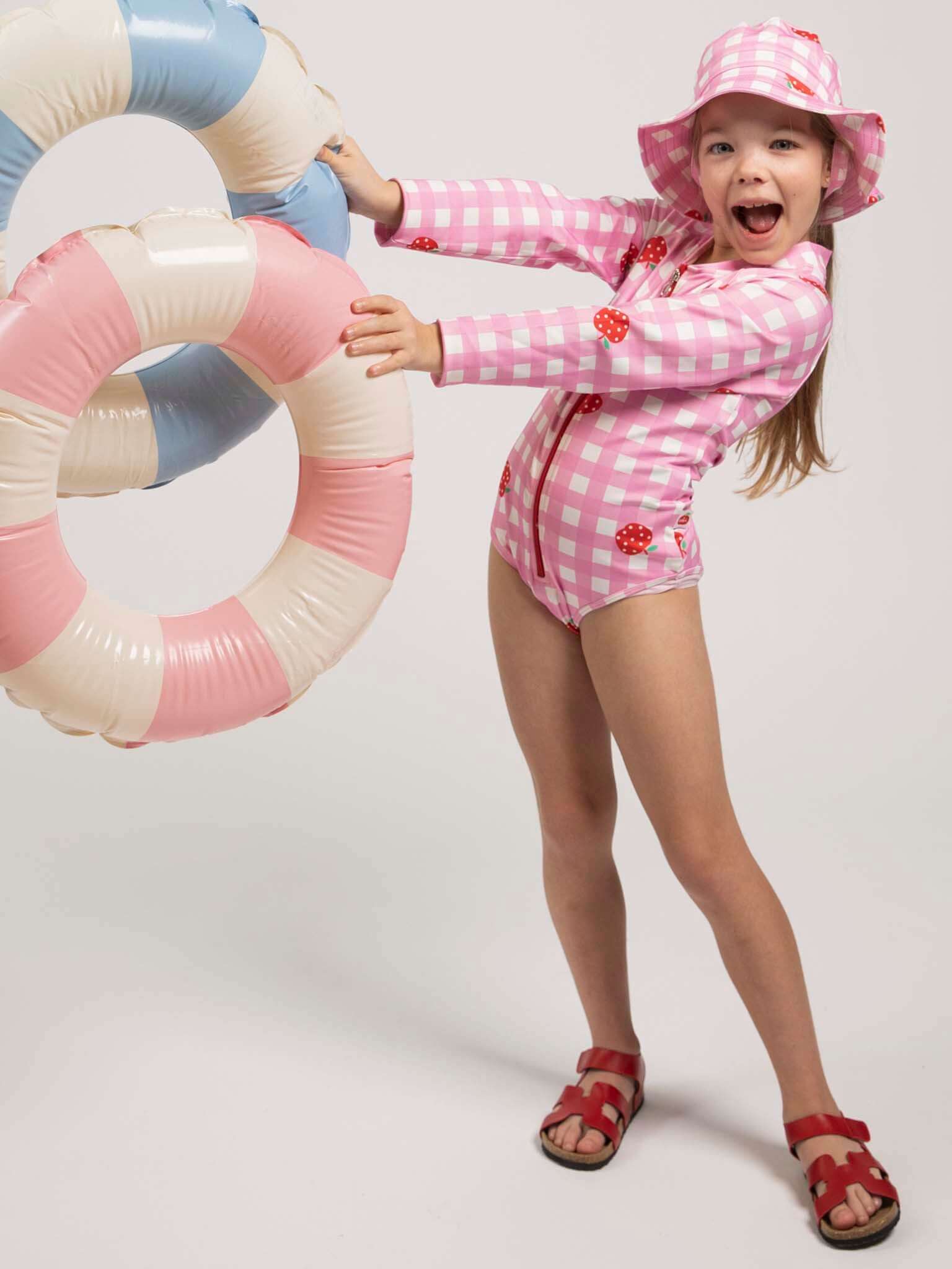 A young girl wearing the Pink Gingham SPF 50 Gingham Swim Ponytail Hat and a pink checkered swimsuit stands on one leg, smiling with her mouth open, holding three pastel-colored swim rings.