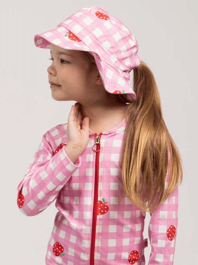 A young girl wears the Pink Gingham SPF 50 Gingham Swim Ponytail Hat, styled with a pink and white checkered outfit featuring strawberry prints, looking to the side with one hand near her neck.