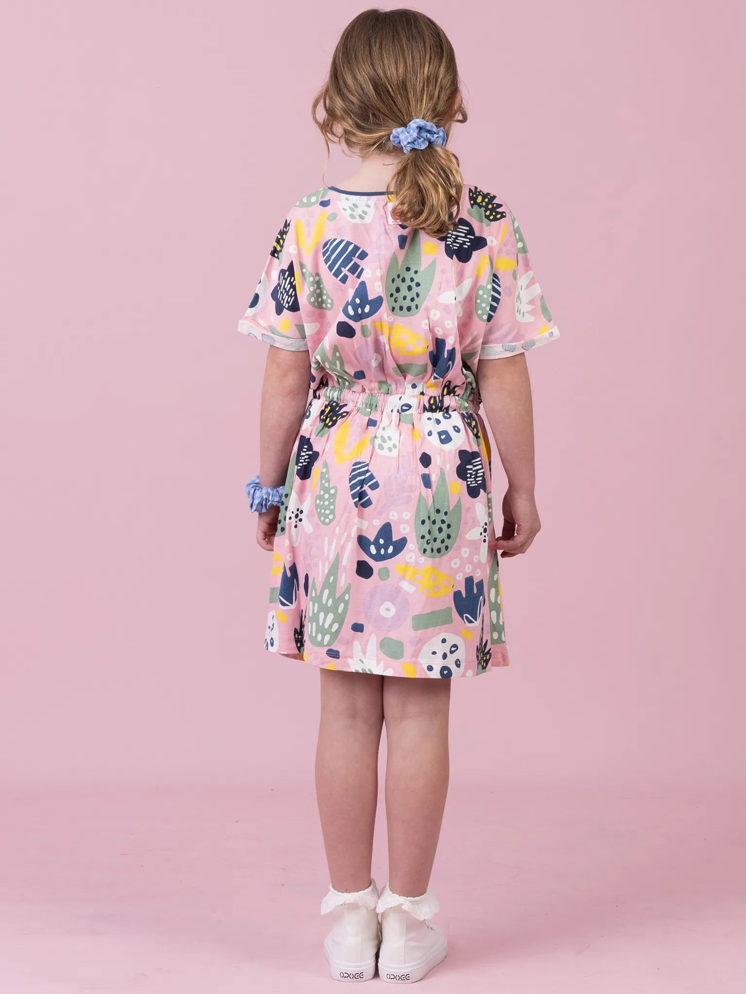 A young girl viewed from behind wearing a short-sleeve pink dress with a colorful abstract botanical pattern, standing against a solid pink background.