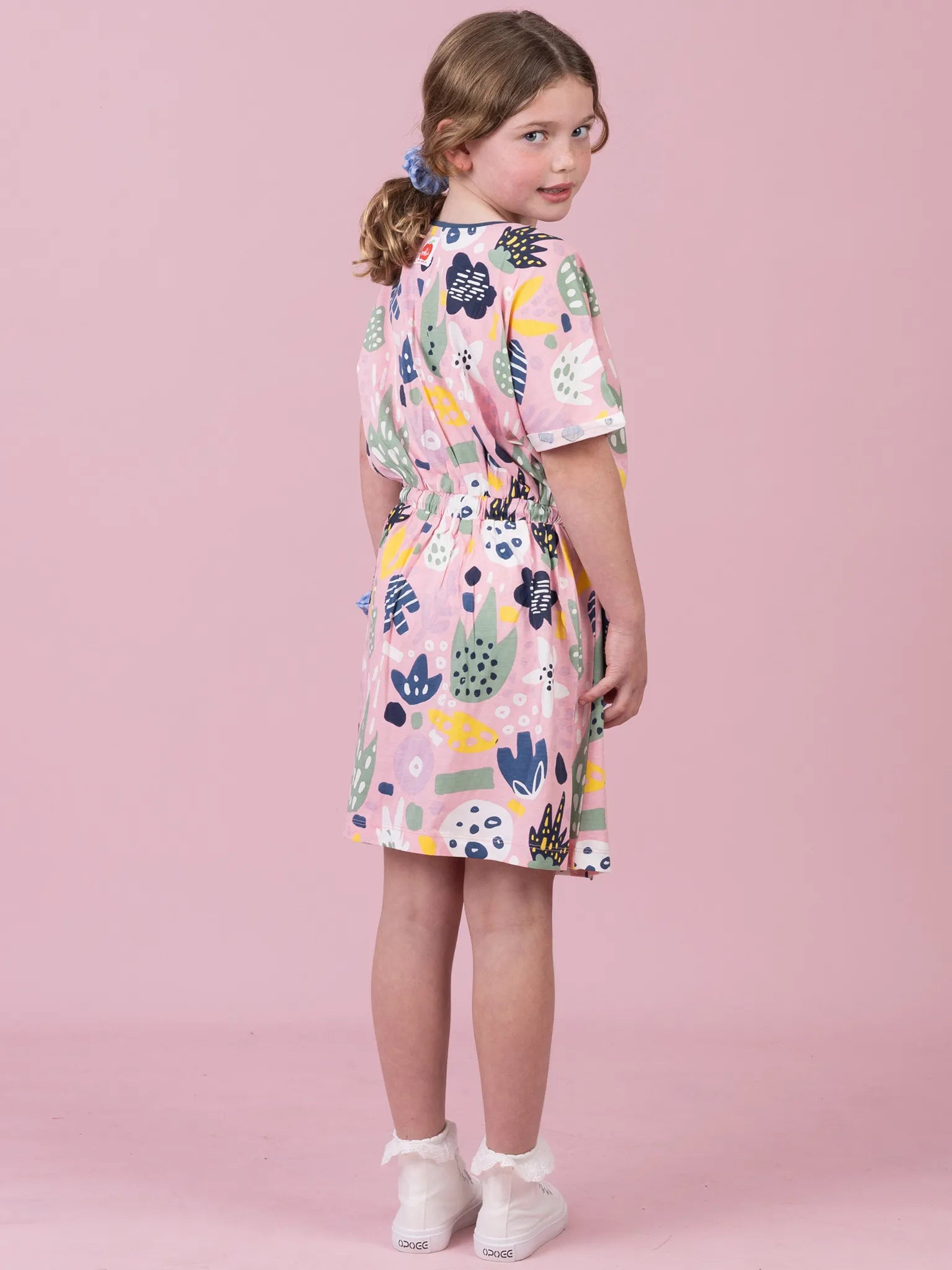 A young girl with her hair in a ponytail is shown from the back wearing a short-sleeved, knee-length dress with a colorful abstract pattern on a white background. She is standing against a solid pink studio backdrop.