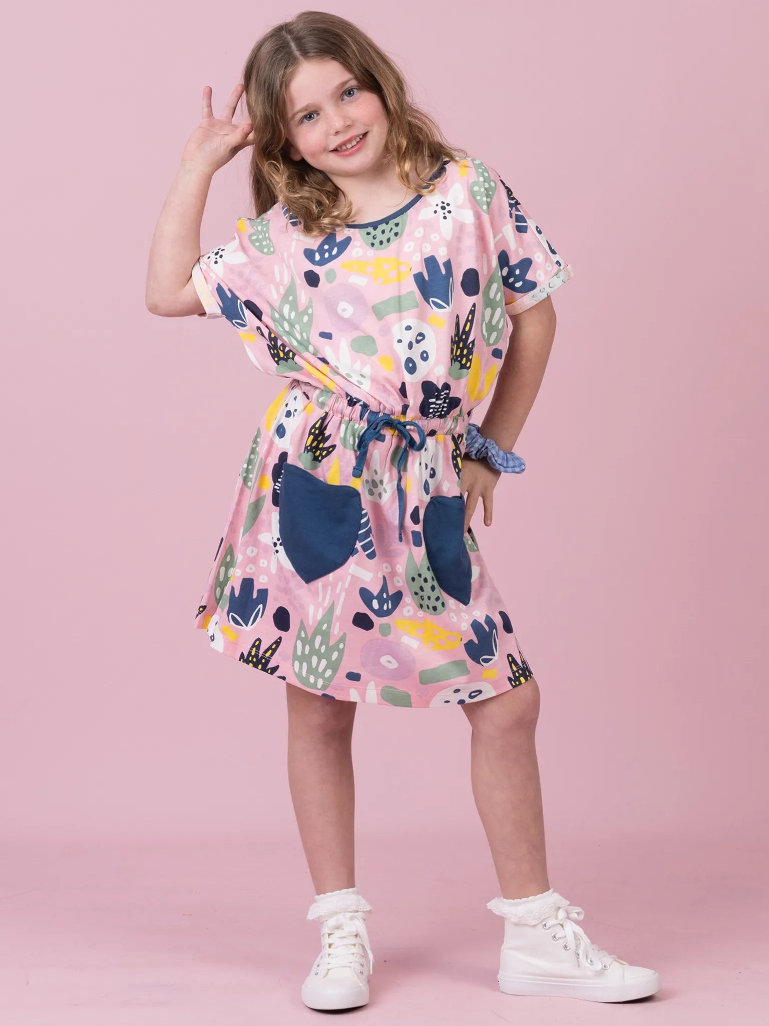 A young girl modeling a short-sleeve pink dress with a colorful abstract print, featuring a tie waist and white sneakers, set against a solid pink studio background.