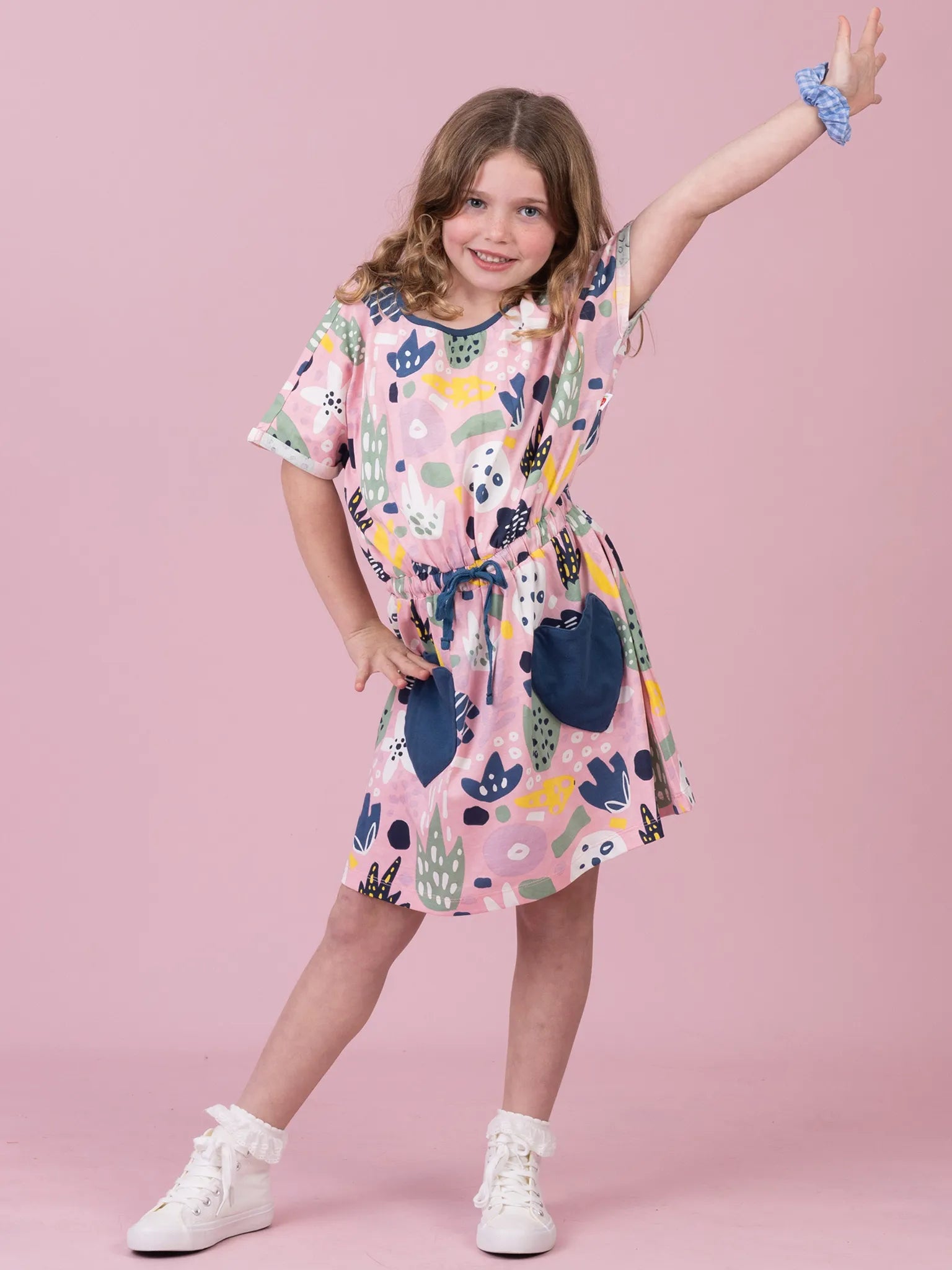 A young girl modeling a short-sleeve pink dress with a colorful abstract pattern and dark blue patch pockets, posed against a solid pink studio background.