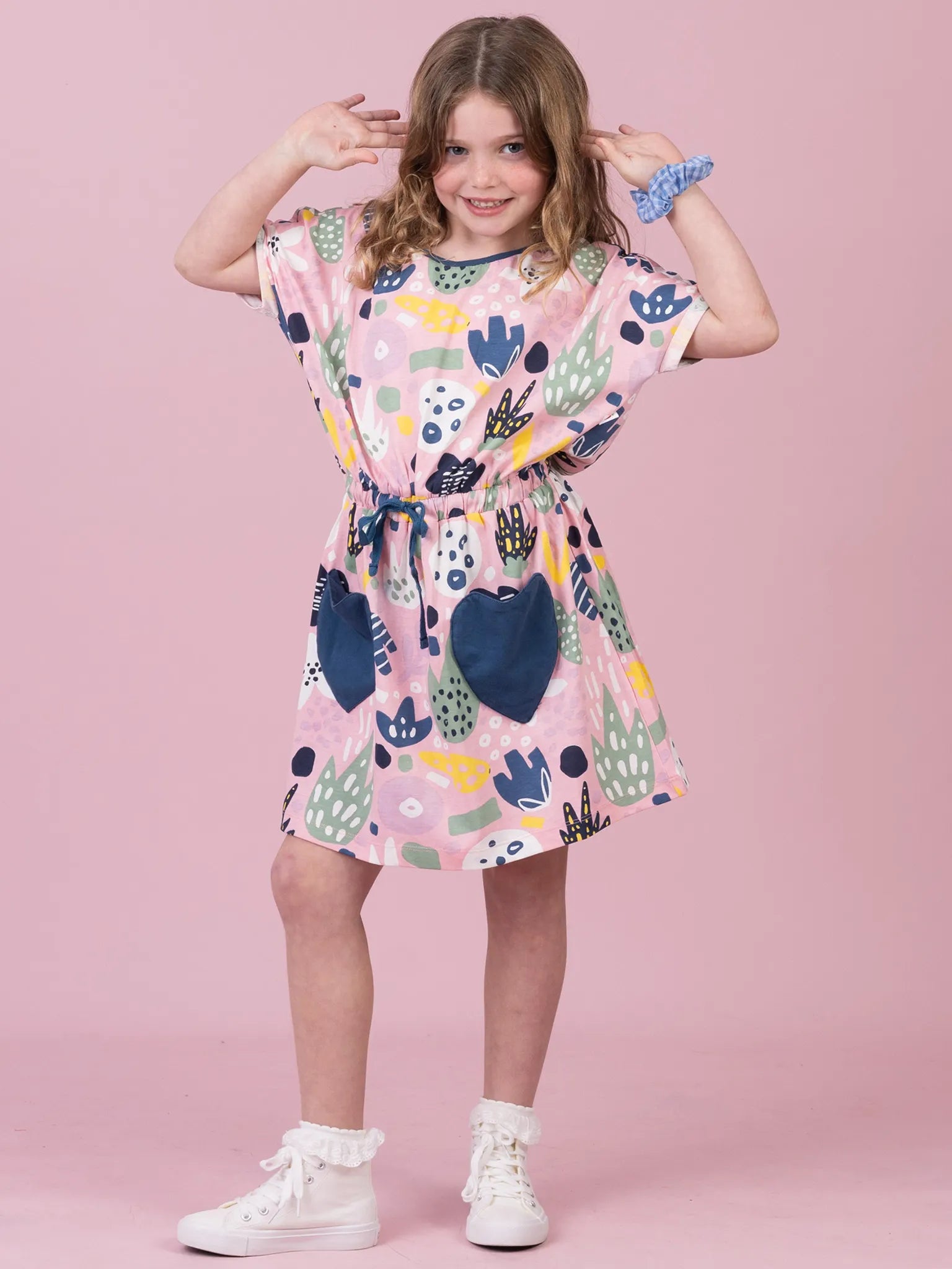 A young girl modeling a short-sleeve pink dress with a colorful abstract pattern, featuring a drawstring waist and white sneakers, set against a solid pink studio background.
