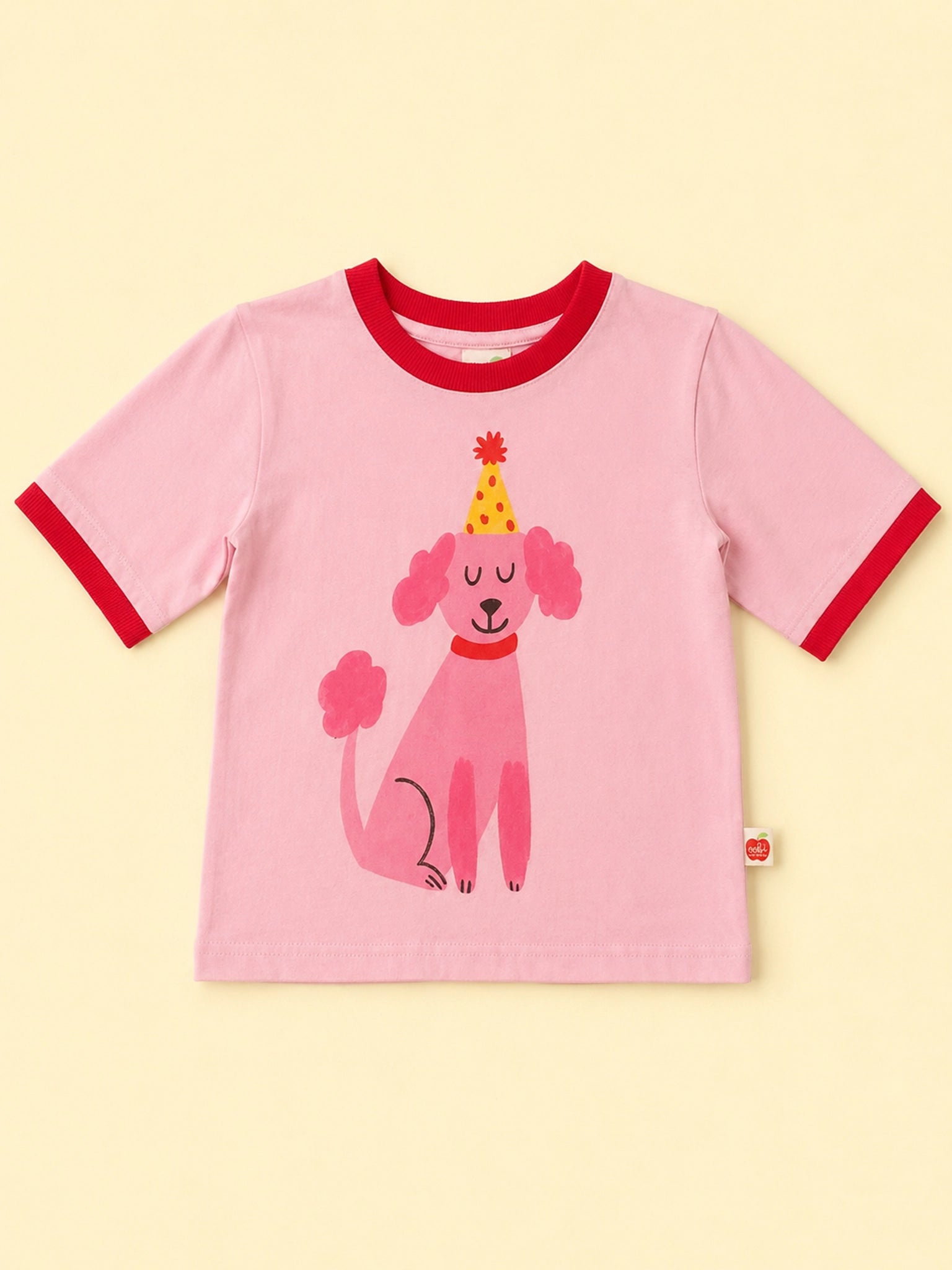 A pink short-sleeve ringer T-shirt with red trim on the collar and cuffs, featuring a cartoon pink poodle wearing a yellow party hat printed on the front, set against a pale yellow background.