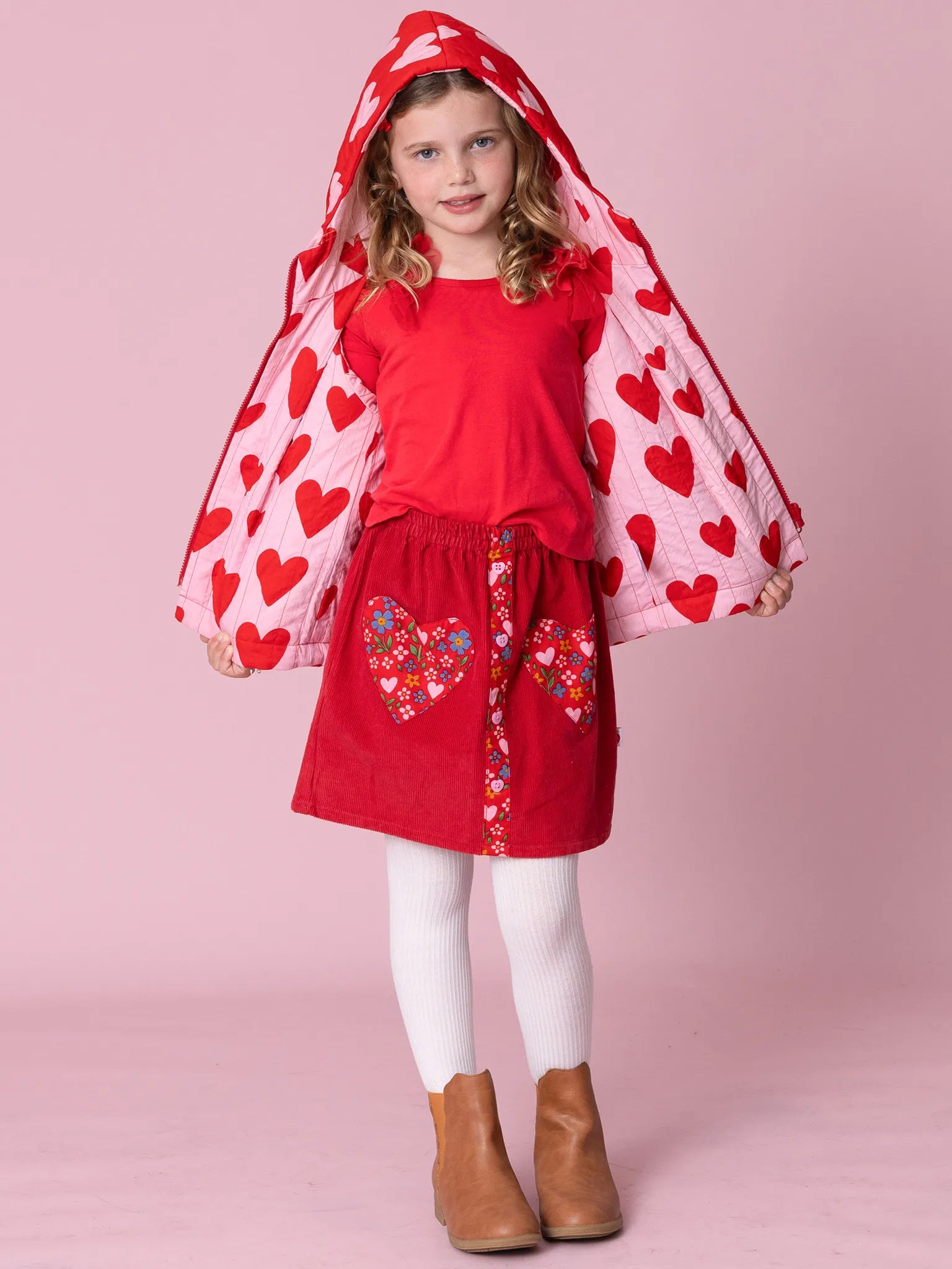 Young girl wearing a red dress with heart patterns and a matching cape on a pink background
