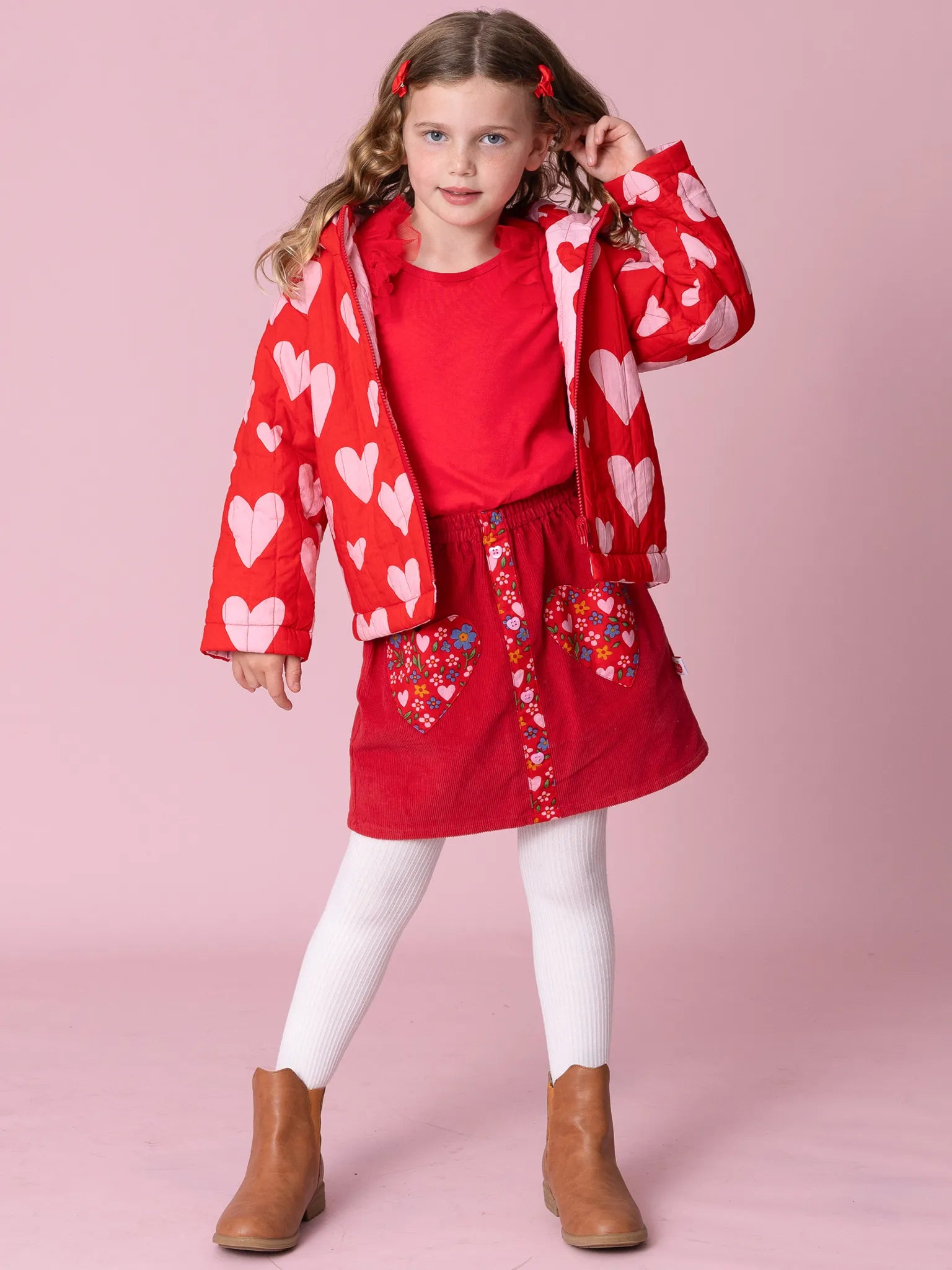 Young girl wearing a red heart-patterned jacket and skirt on a pink background