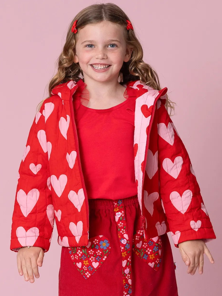 Young girl wearing a red heart-patterned jacket against a pink background