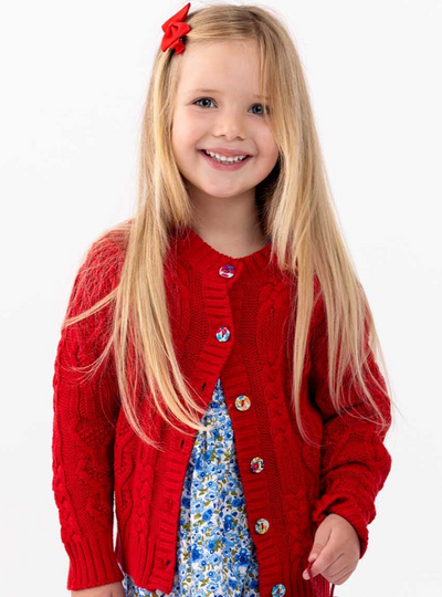 A young girl with long blonde hair smiling, wearing a Willow Deep Red Cotton Cardigan by Knitwear and a blue floral dress, accented with a red bow clip in her hair.