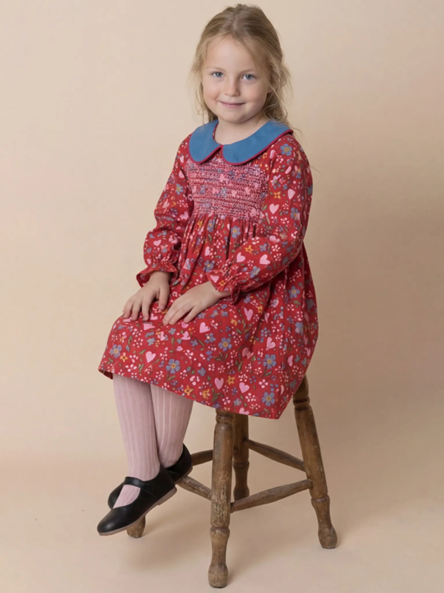 Young girl wearing a red floral dress with a blue collar, sitting on a wooden stool against a beige background.
