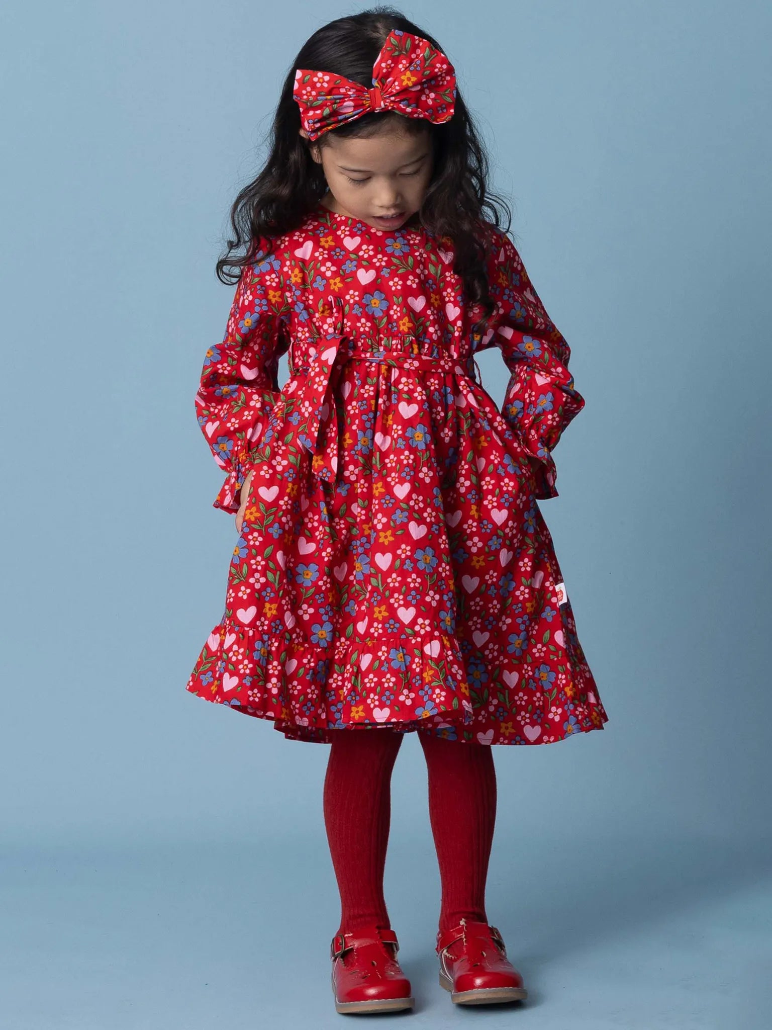 Young girl wearing a red floral dress with matching headband and shoes on a blue background
