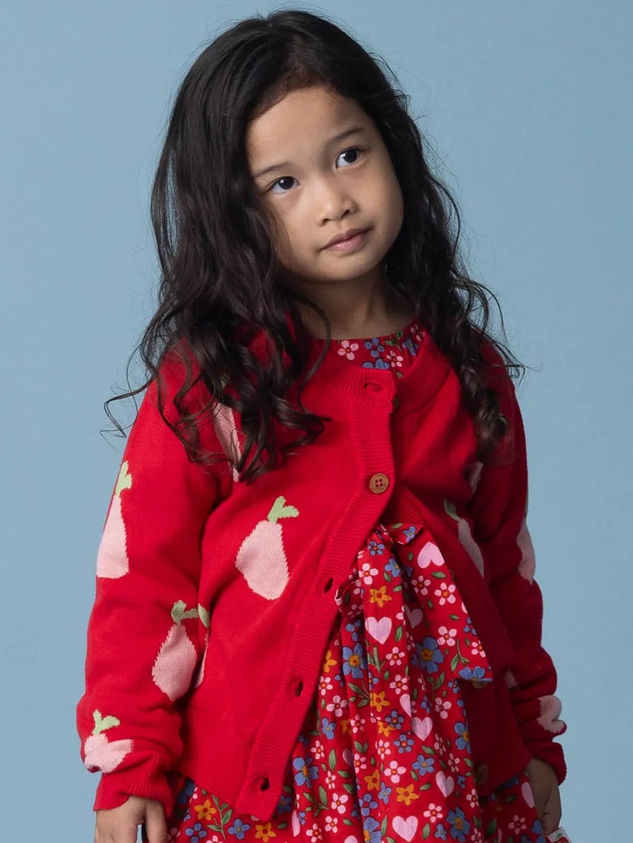 Young girl wearing a red cardigan with heart patterns and a colorful dress against a light blue background