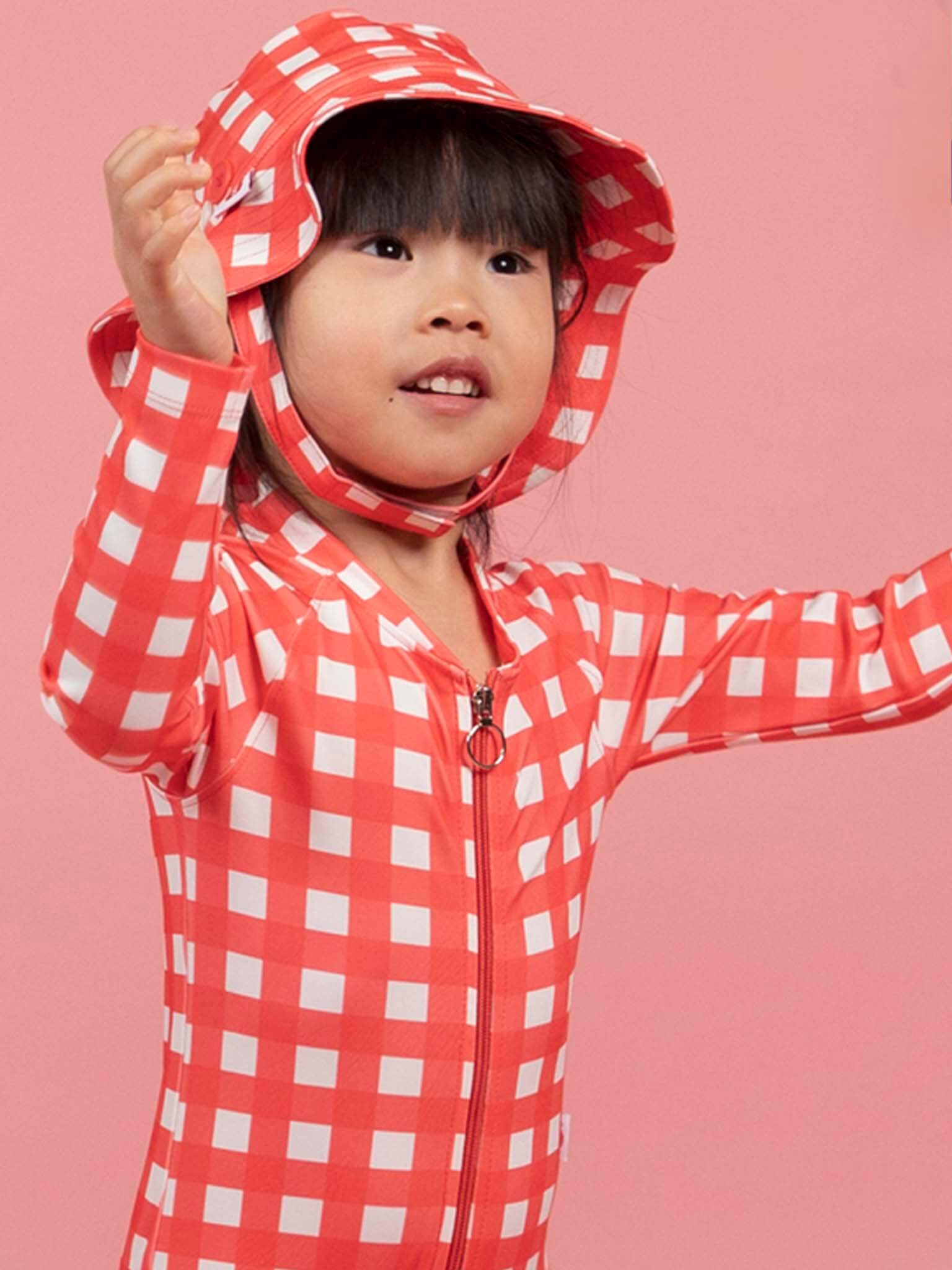 A young child wearing the Red Gingham SPF 50 Gingham Swim Ponytail Hat stands against a pink background with arms raised, paired with a matching zip-up outfit.