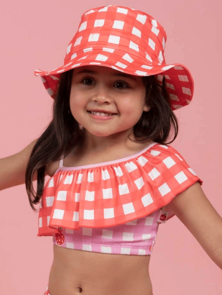 A young girl smiles in a Red Gingham SPF 50 Gingham Swim Ponytail Hat with a matching off-shoulder top, standing against a pink background.