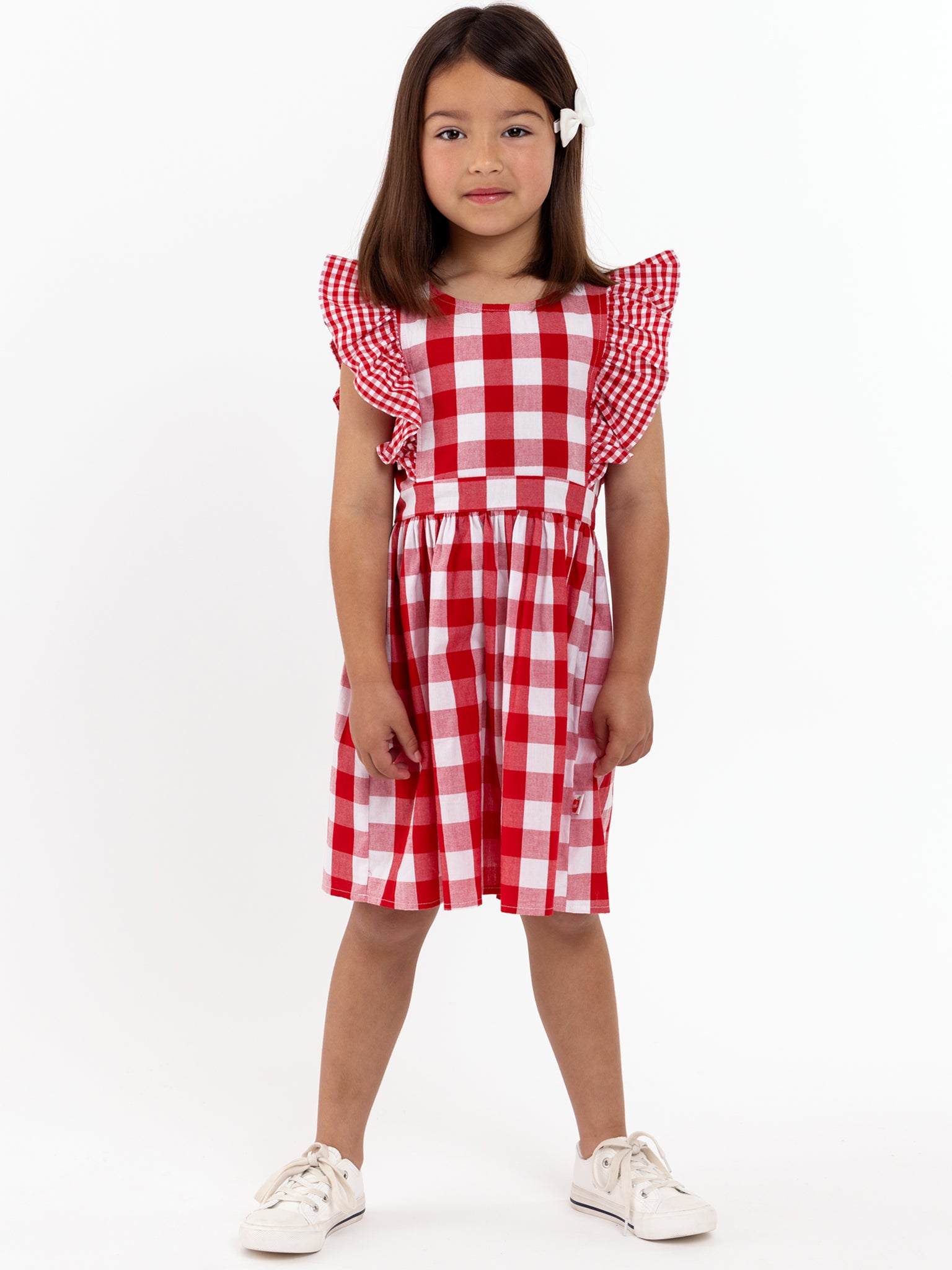 A young girl stands facing forward in a Red Check Jayne Dress by Red Check, featuring ruffled sleeves, paired with white sneakers and a white bow, set against a plain white background.