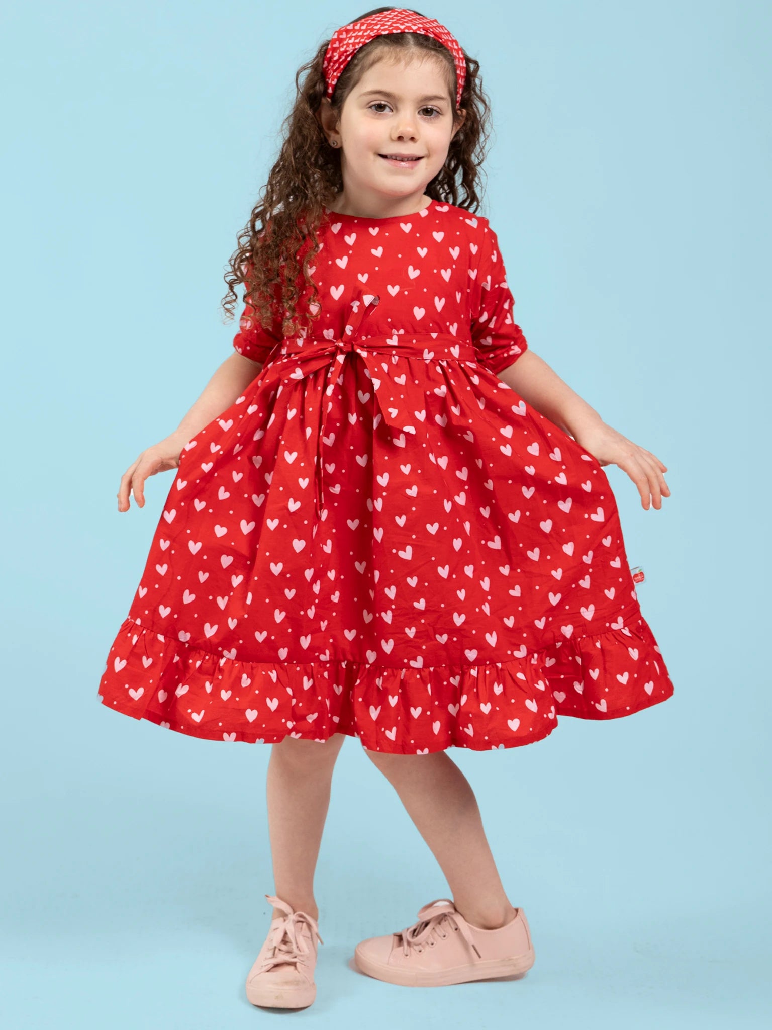 A young girl poses against a blue background in the Hearts Red & Pink Heart Midi Dress, crafted from ethically made cotton, paired with pink shoes and a red headband.