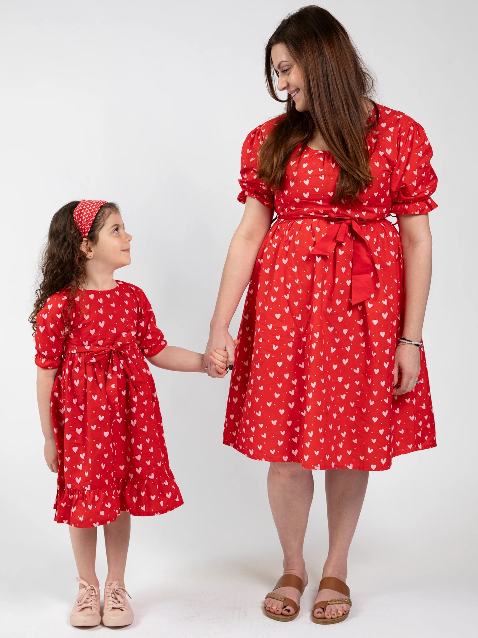 An adult and child smile, holding hands in matching Hearts brand Red & Pink Heart Midi Dresses. Ethically made, these dresses feature playful white heart patterns for a joyful, coordinated look.