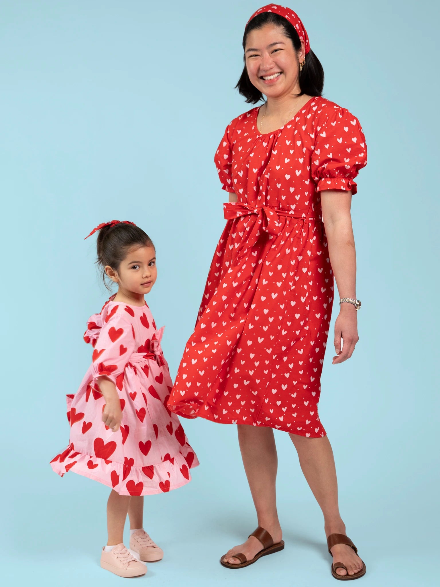 An adult and a child wear matching red and white heart-print dresses with Essentials Red and Pink Hearts Headbands, posing together against a light blue background.