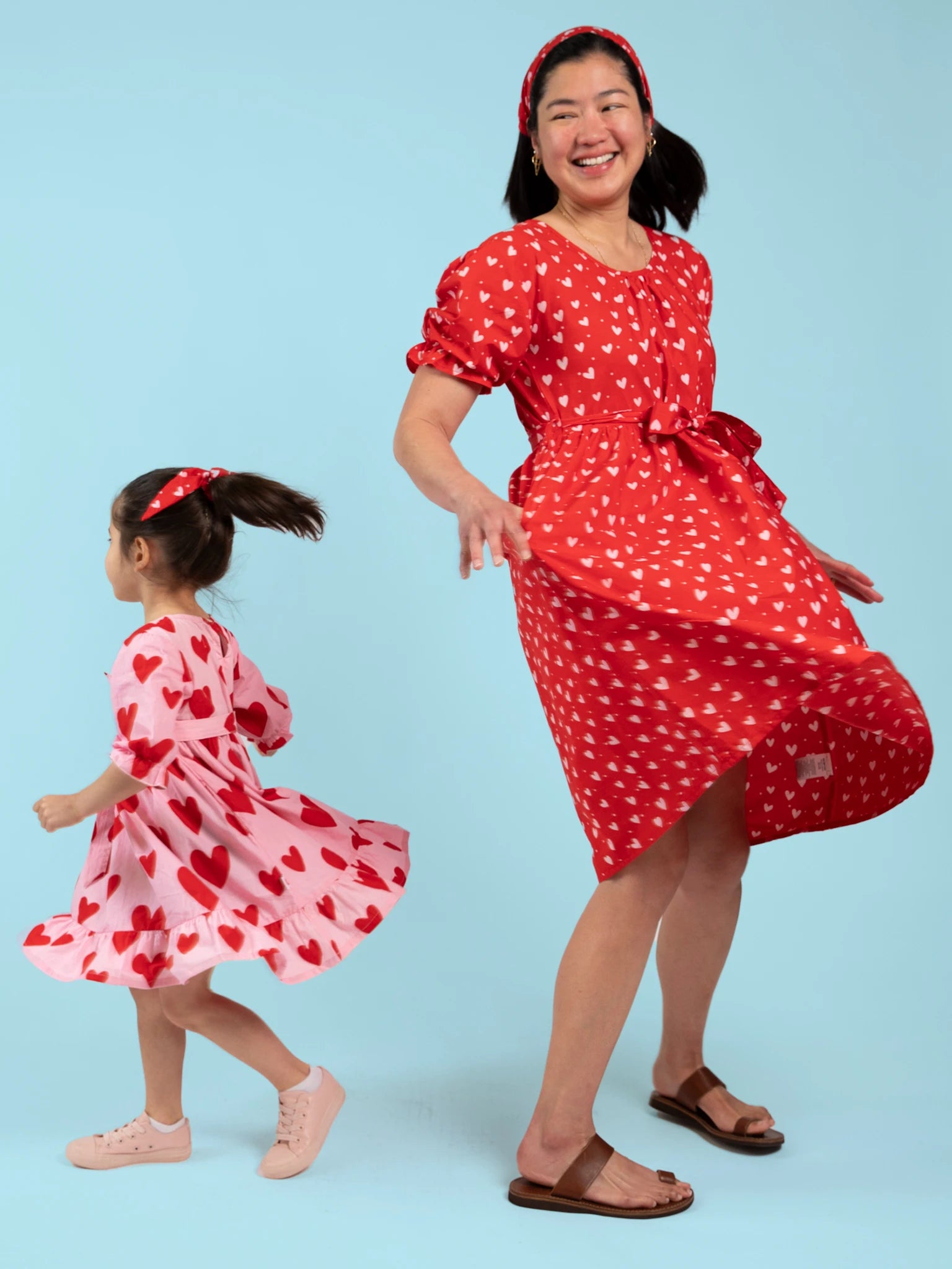 A woman and child wear matching Hearts Pink & Red Heart Women's Midi Dresses and headbands, featuring hand-printed heart patterns, as they pose playfully against a light blue background.