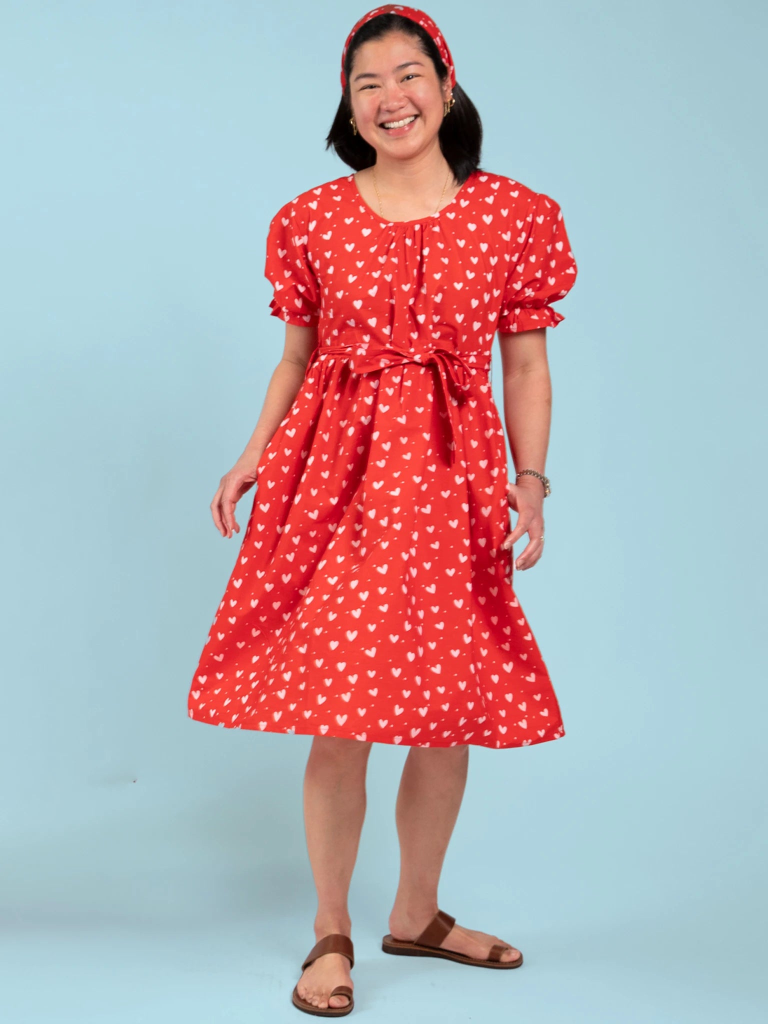 A woman stands and smiles against a light blue background, wearing an Essentials Red and Pink Hearts Headband with a red dress covered in hand-printed hearts and brown sandals.